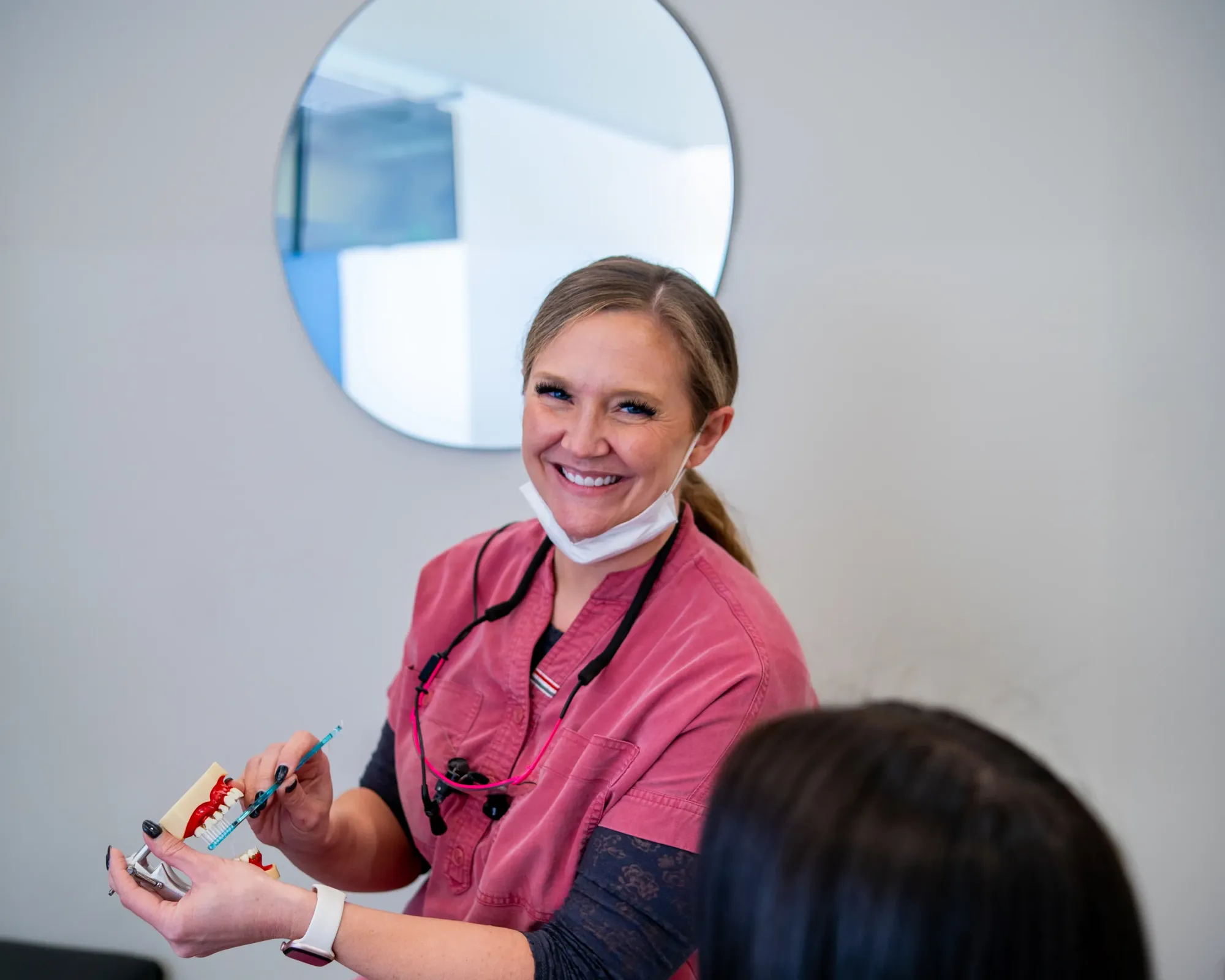 Smiling female dental professional in pink scrubs demonstrating teeth cleaning on a dental model.