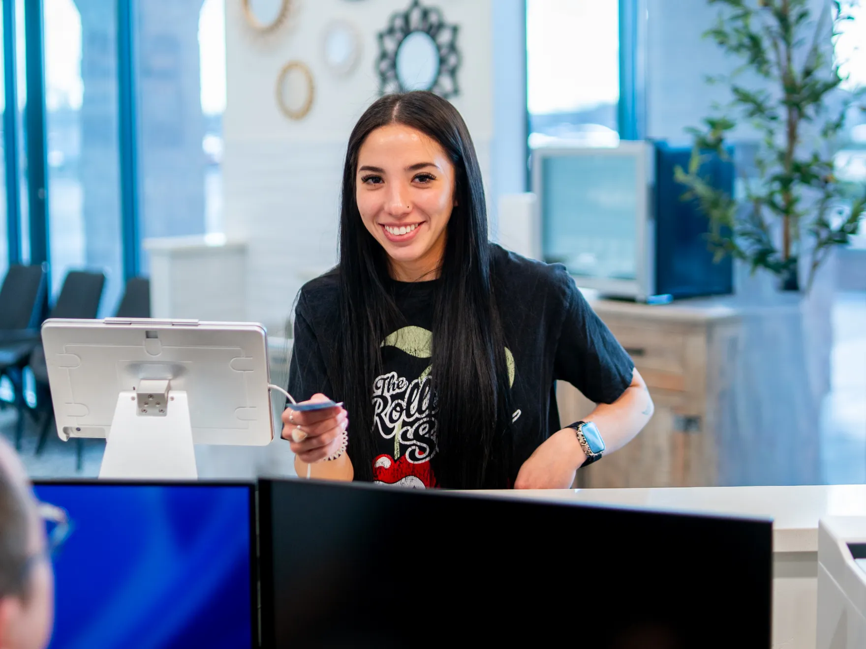 Smiling woman with long dark hair standing behind a reception desk holding a card near a tablet screen.