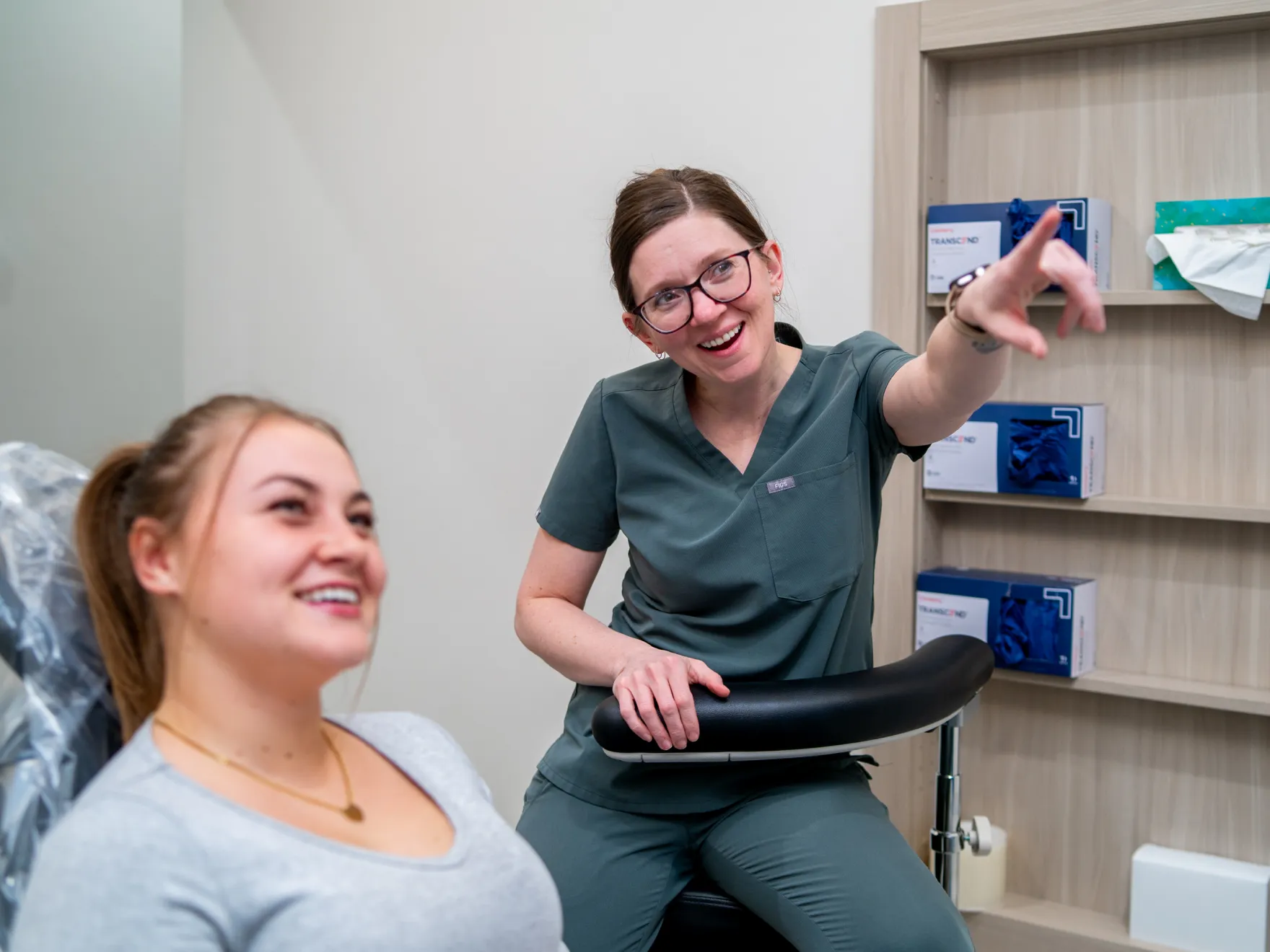 Female healthcare professional in green scrubs smiling and pointing while talking to a patient seated in a medical chair.