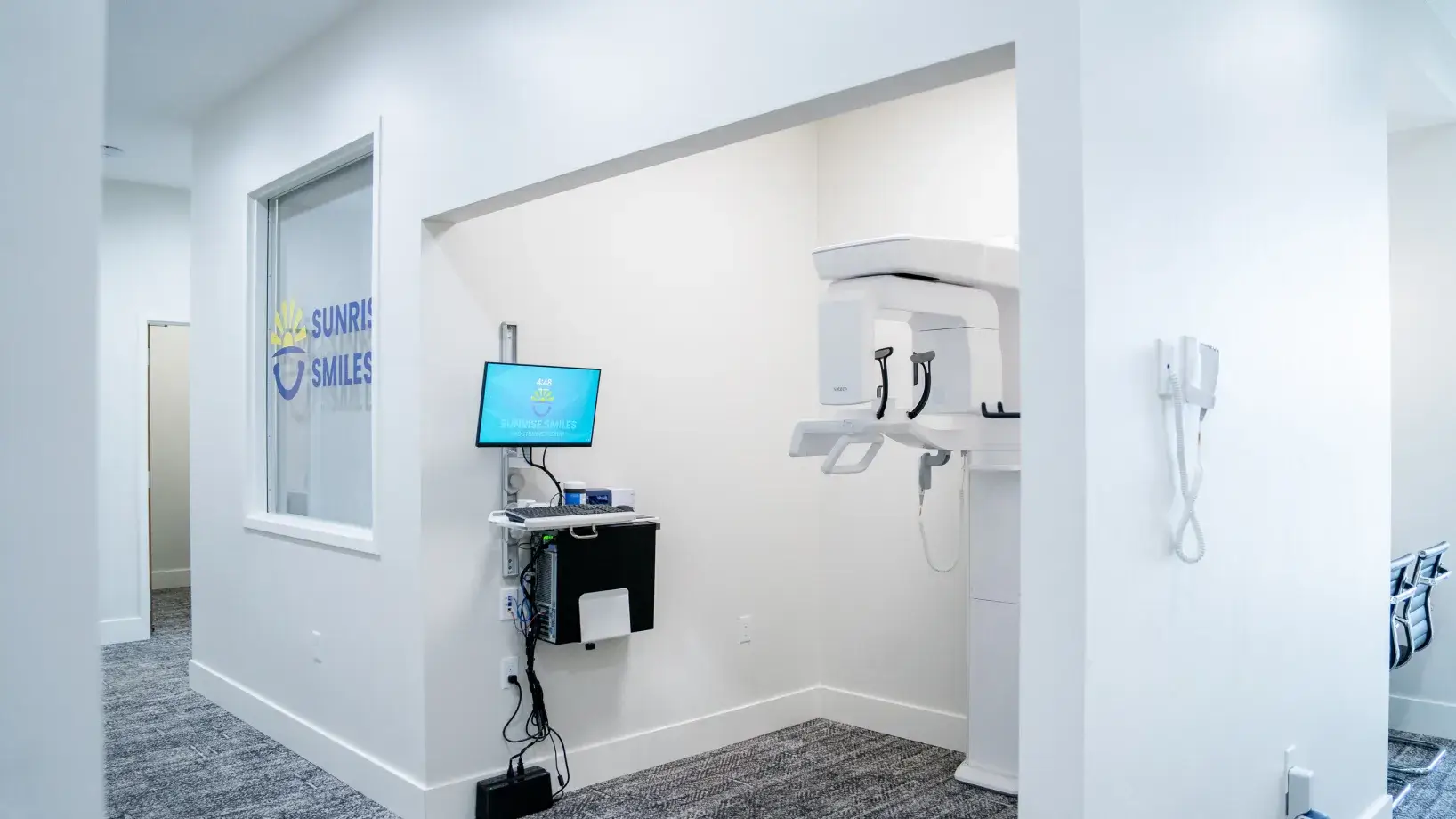Modern dental X-ray machine in a white-walled clinic room with a computer monitor on the side.