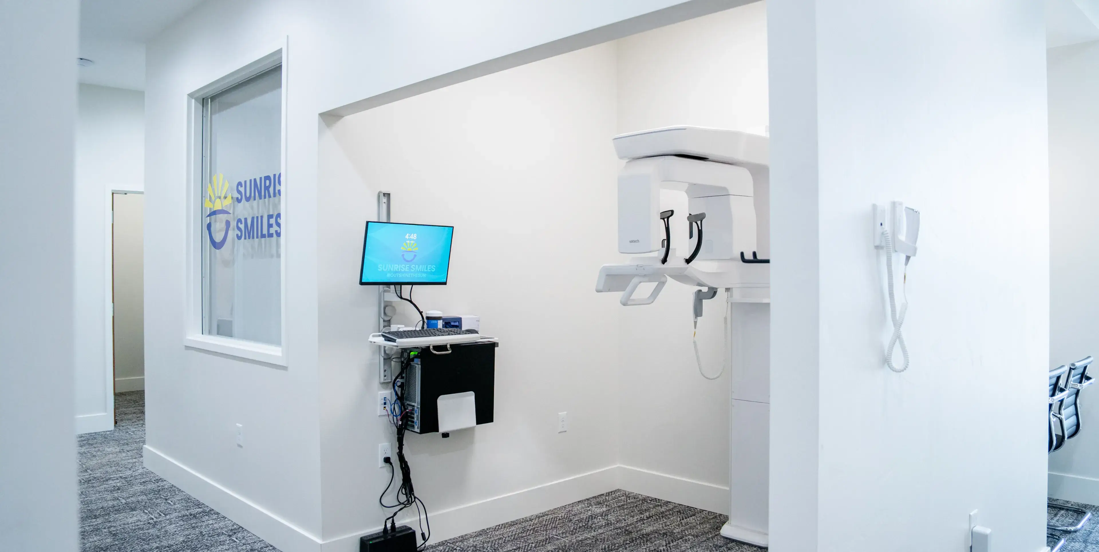 Modern dental office corner with a panoramic dental X-ray machine and wall-mounted computer screen showing Sunrise Smiles logo.