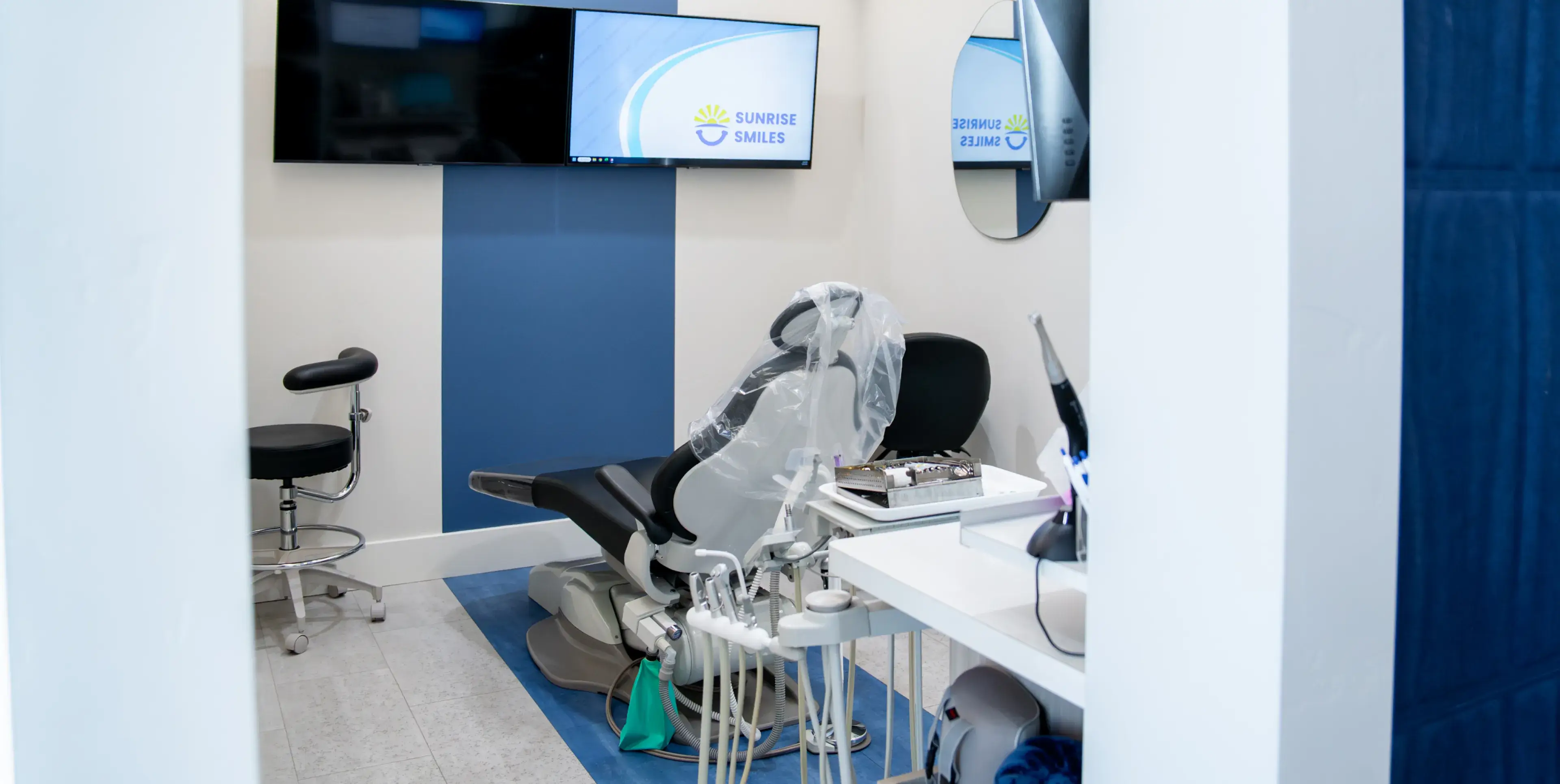 Modern dental exam room featuring a dental chair covered in plastic, dental instruments on a tray, a black stool, and a wall-mounted TV displaying the Sunrise Smiles logo.