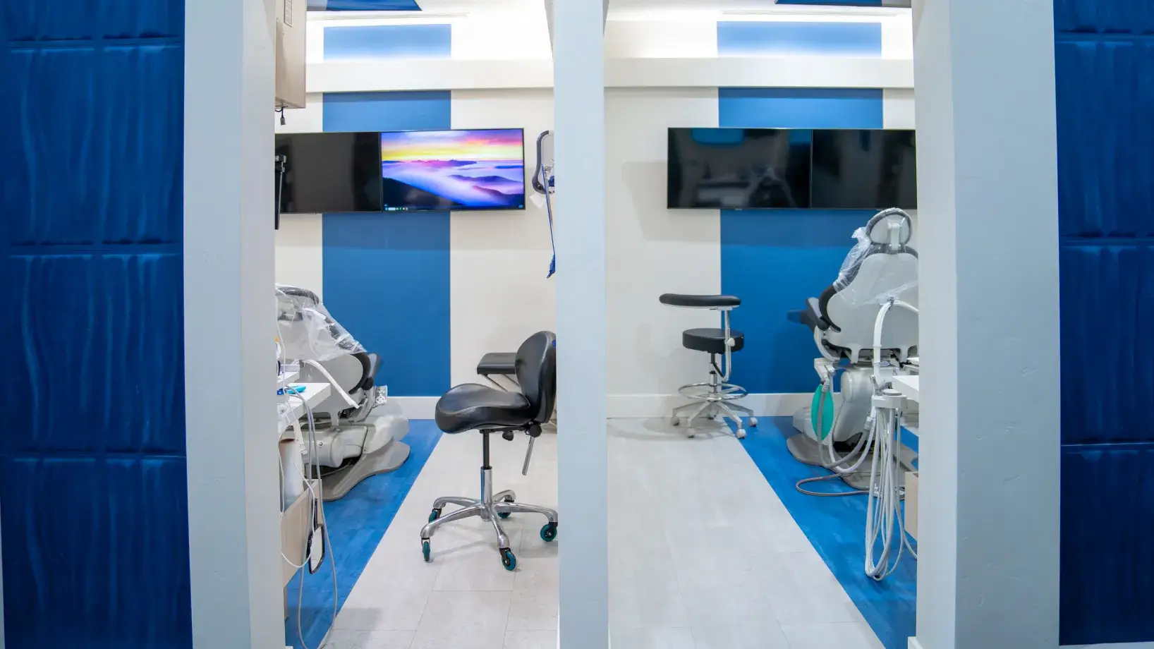 Modern dental clinic room with two dental chairs separated by a partition and mounted flat-screen TVs on blue and white walls.