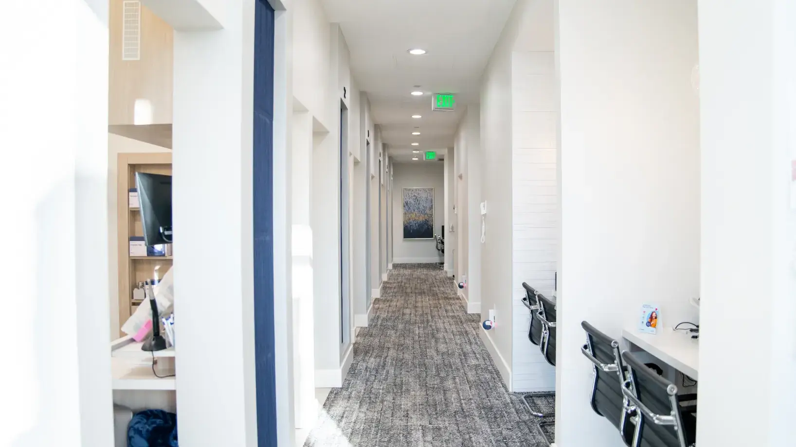Bright modern office hallway with gray carpet, white walls, and black chairs along desks on the right side.