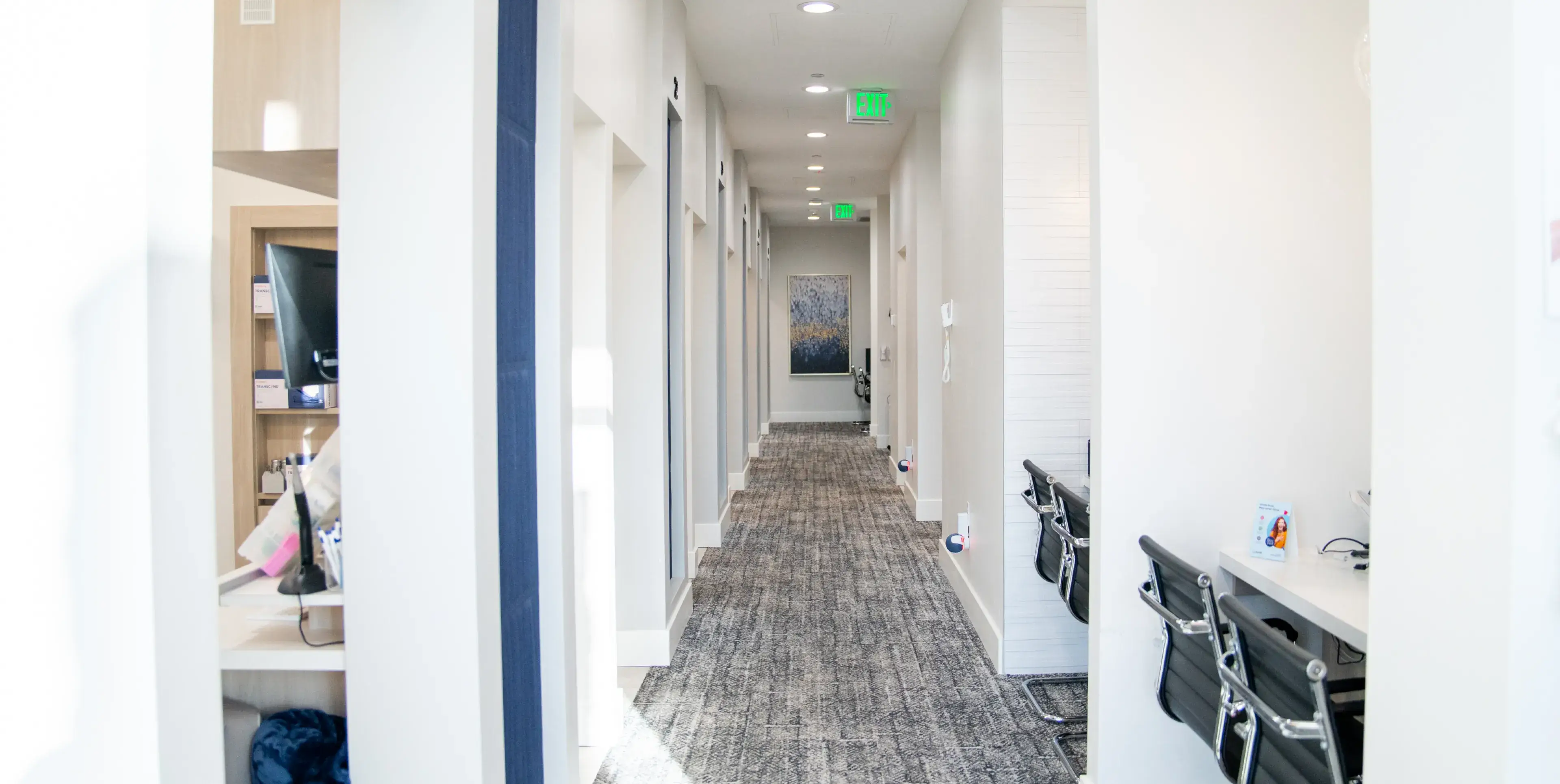 Modern office hallway with grey carpet, white walls, black chairs at workstations, and exit signs overhead.