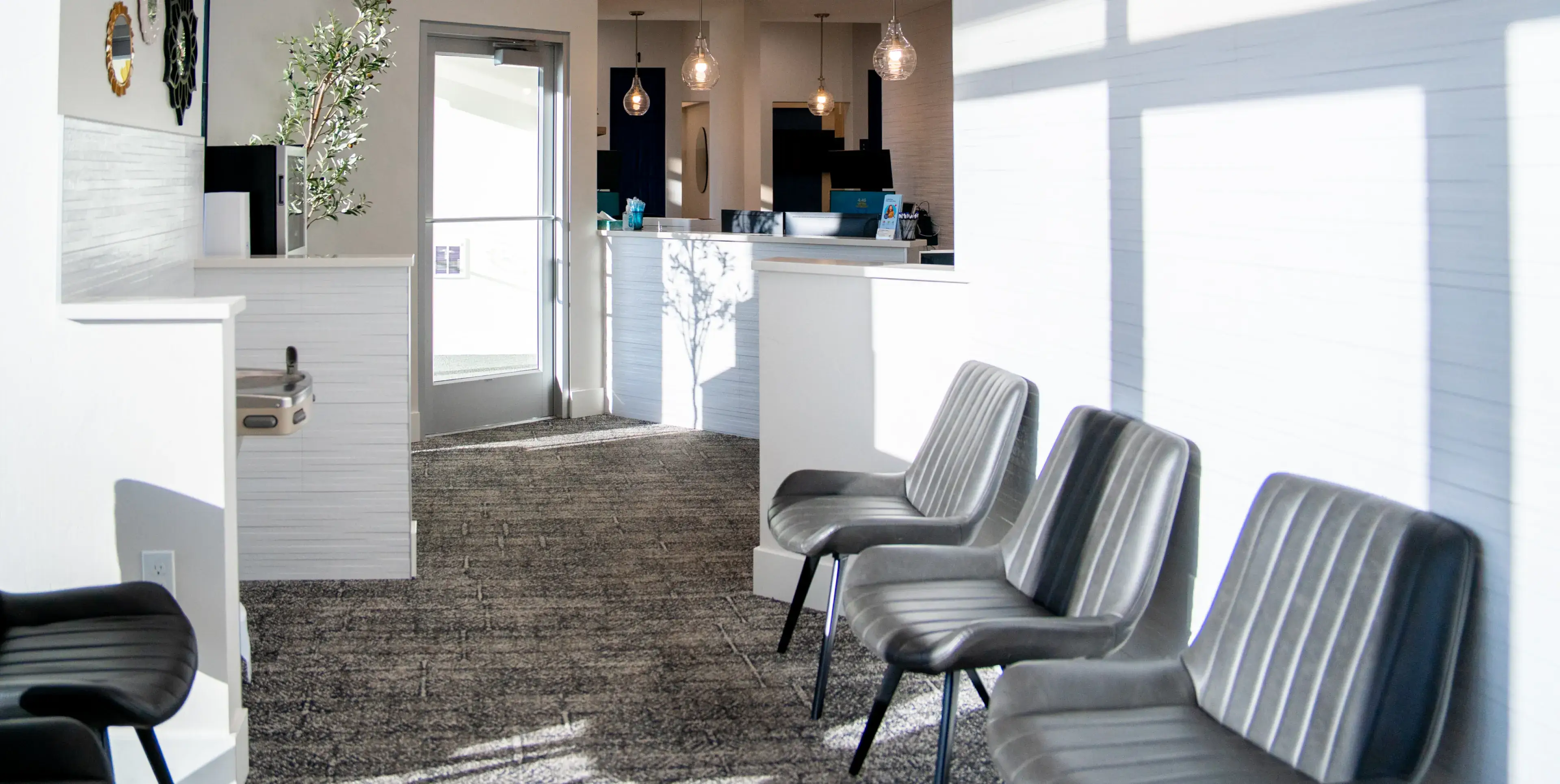 Modern office waiting area with three gray chairs, a reception desk, hanging pendant lights, and sunlight casting shadows on white walls.