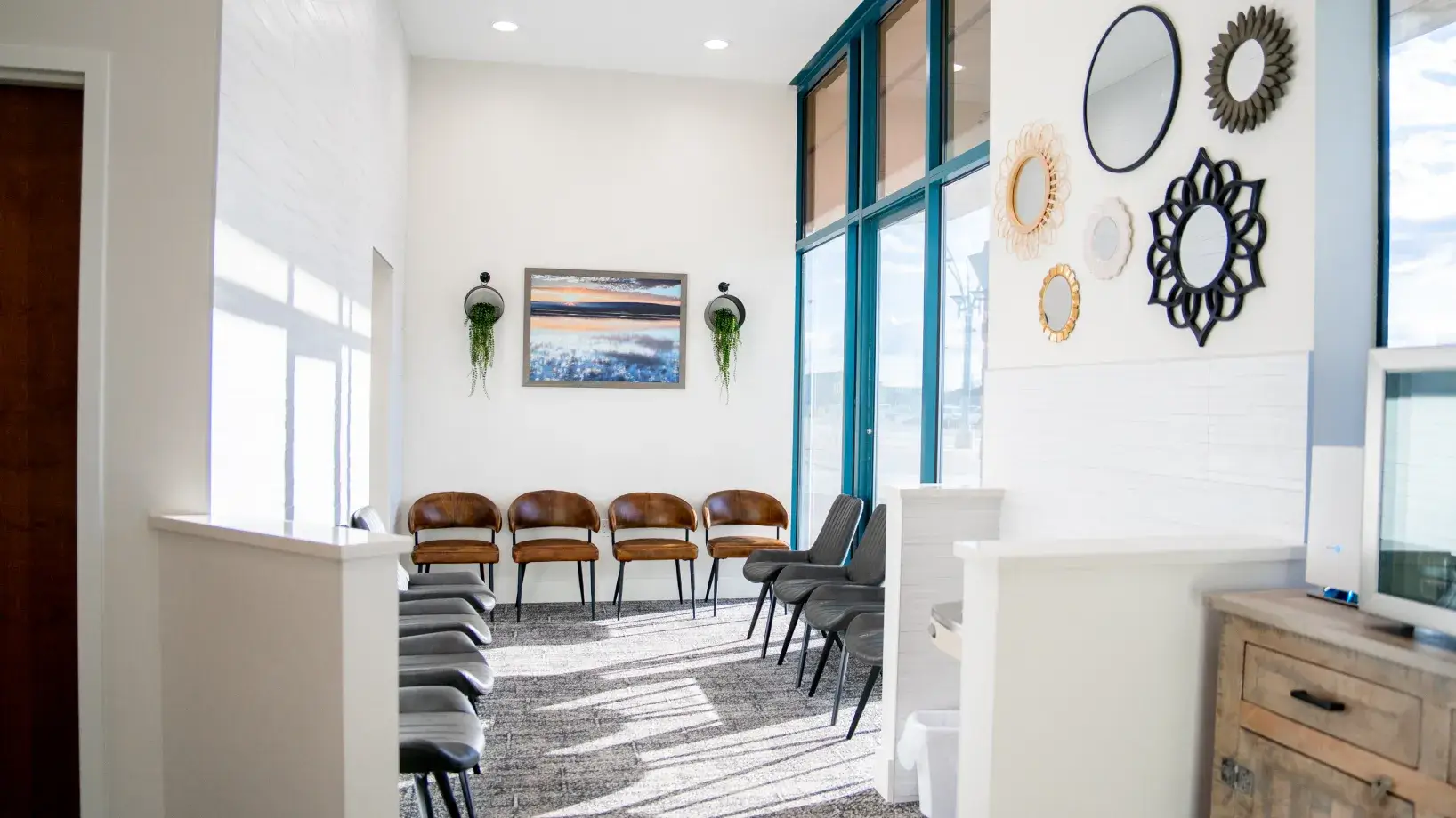 Bright waiting room with brown and gray chairs arranged along white walls, large windows, decorative mirrors, and a painting with hanging plants.