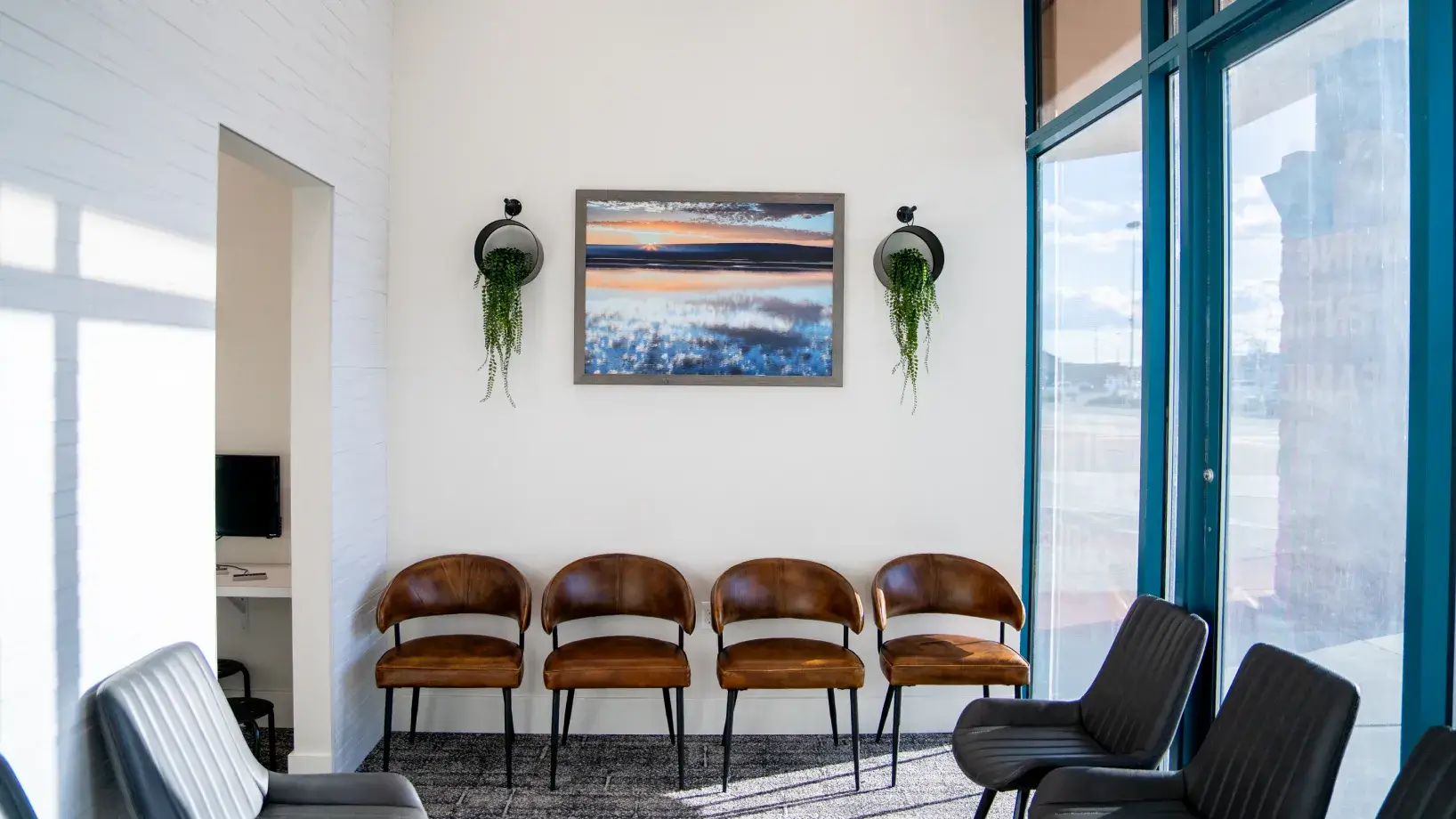 Waiting room with four brown leather chairs in a row, two black chairs nearby, a framed landscape photo, and two hanging plants on a white wall.