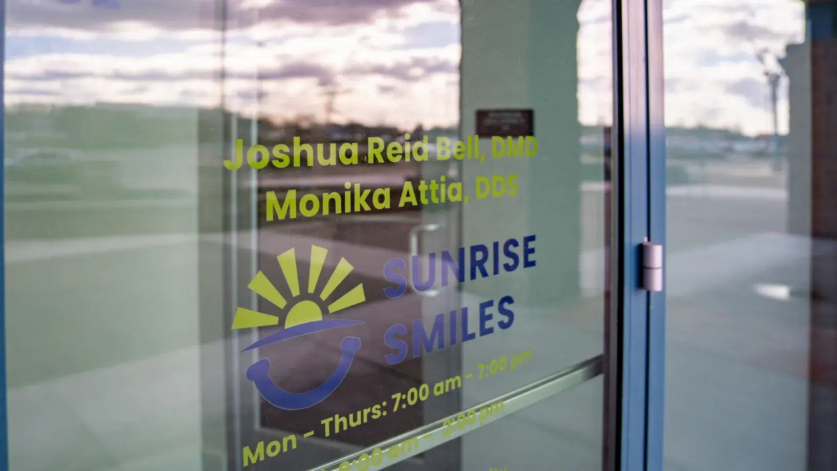 Glass door of Sunrise Smiles dental office showing names Joshua Reid Bell, DMD and Monika Attia, DDS with business hours.