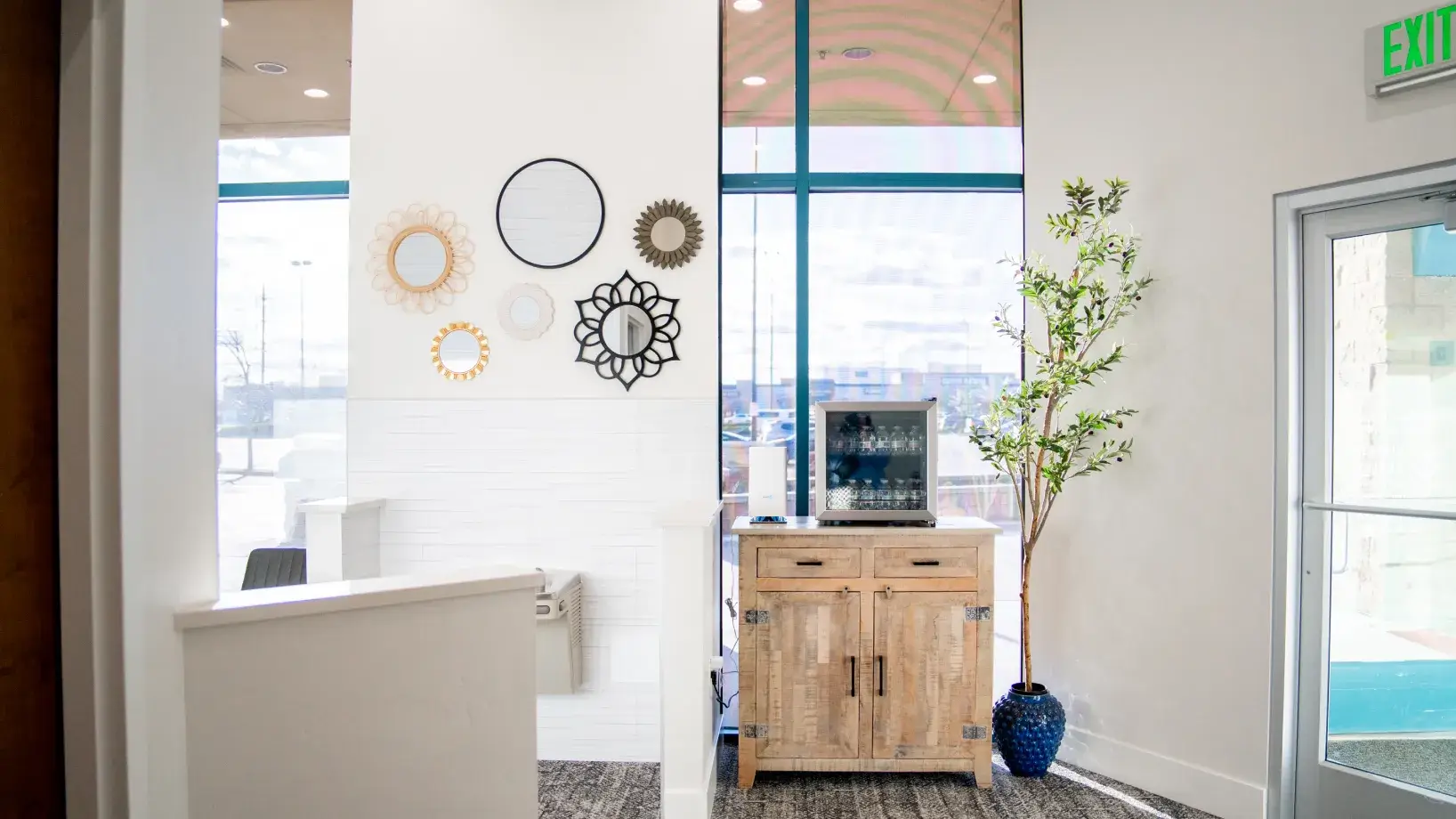 Bright waiting area with a wooden cabinet holding a water dispenser, decorative round mirrors on the wall, and a tall plant in a blue pot beside an exit door.