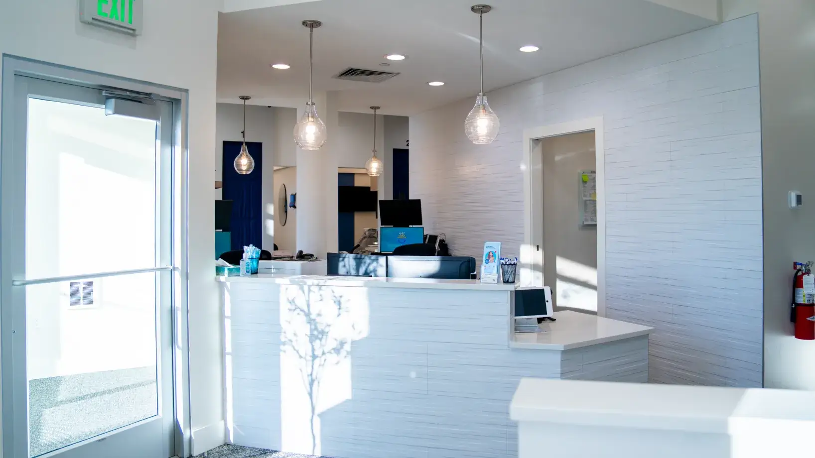 Bright modern reception area with white desk, hanging pendant lights, computer monitors, and an exit door to the left.