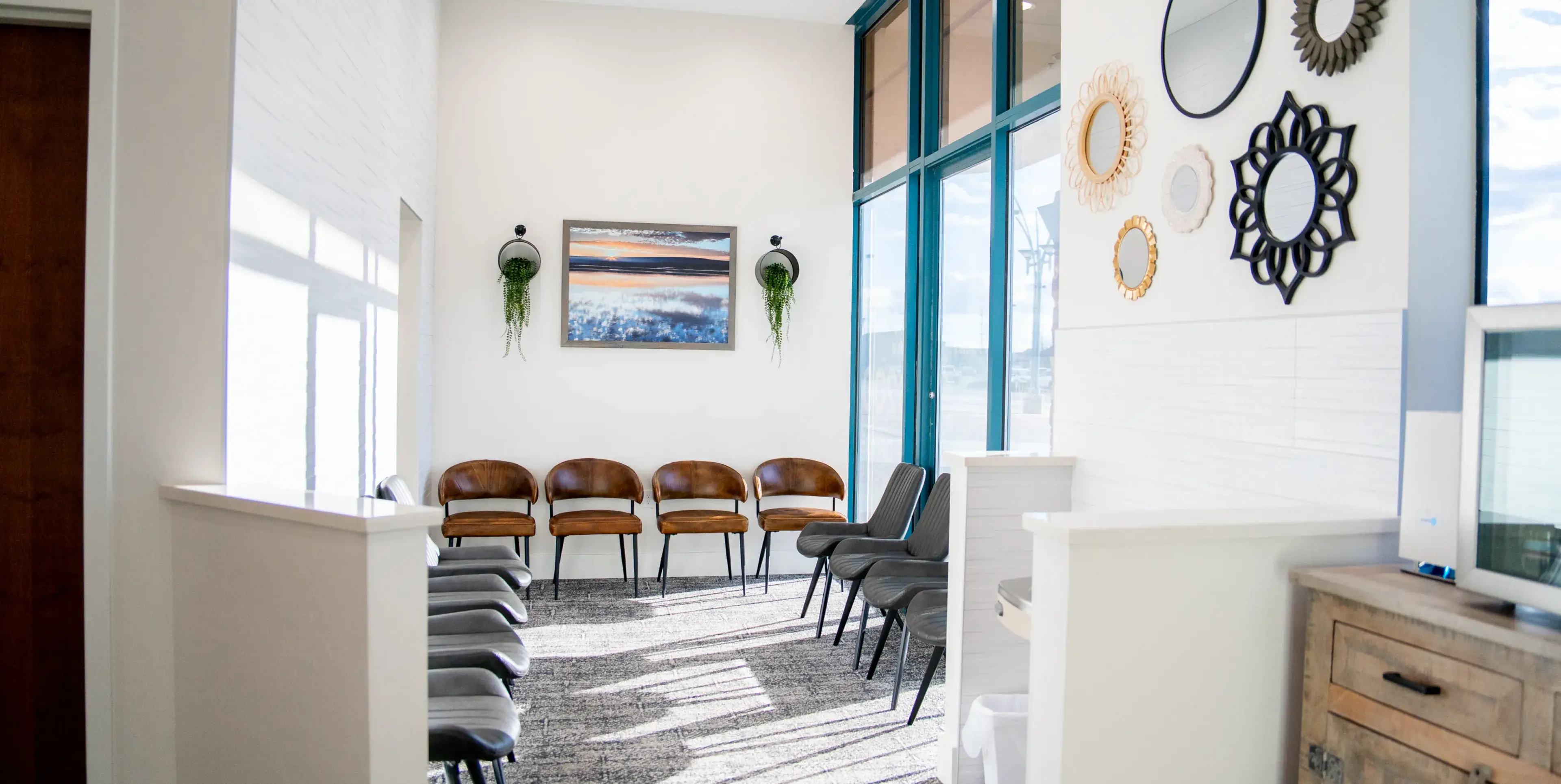 Bright waiting room with brown and gray chairs, large windows, wall mirrors, and framed landscape artwork.