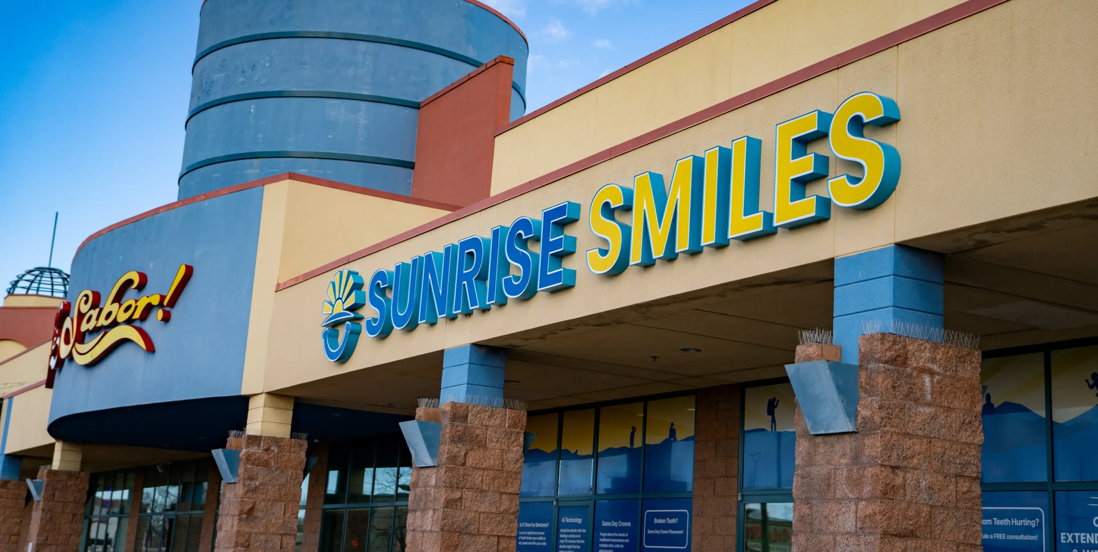 Exterior view of Sunrise Smiles dental office with a blue and yellow sign on a beige and blue building.