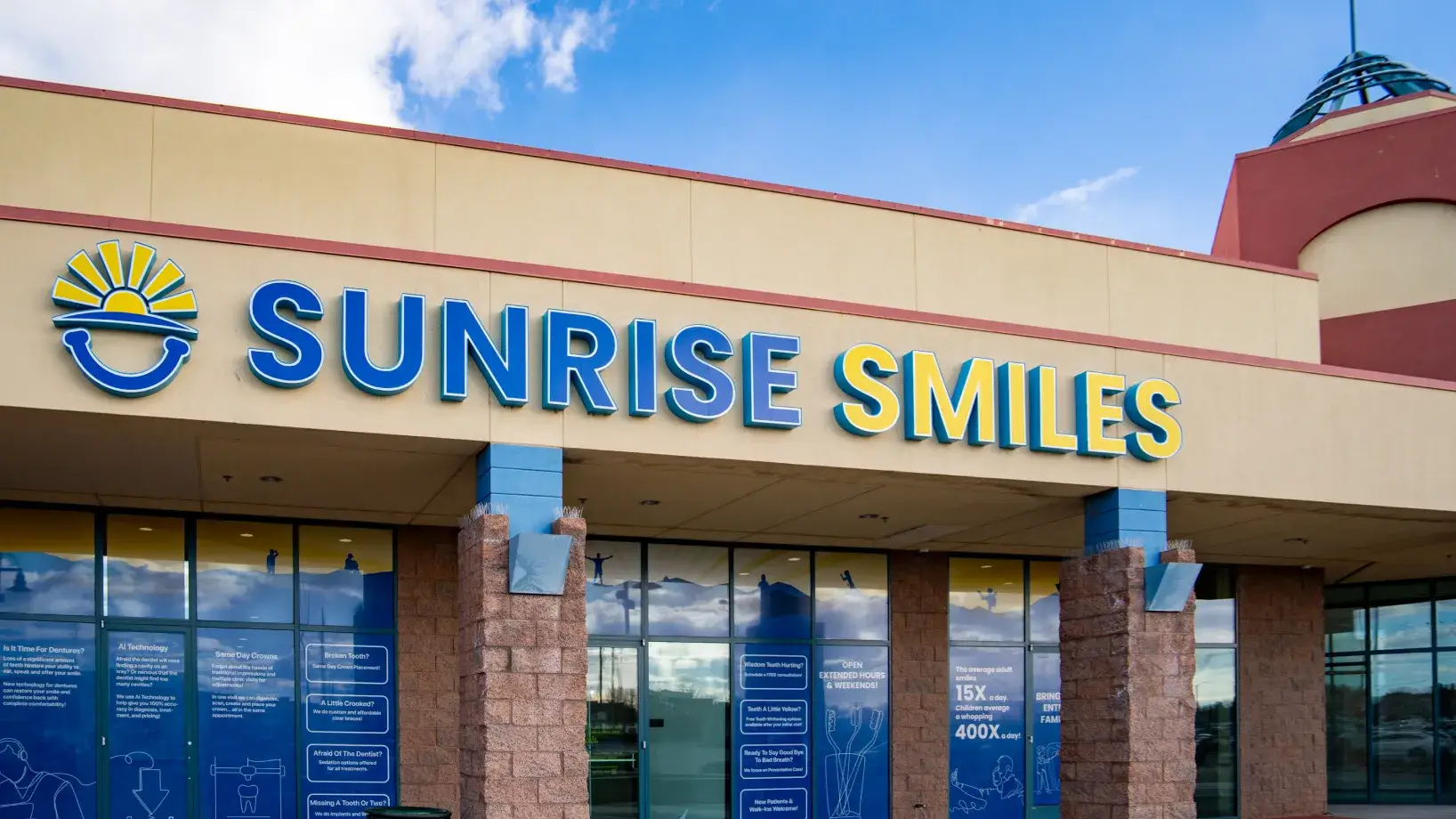 Exterior view of Sunrise Smiles dental office with large blue and yellow signage and sunlight reflected on windows.