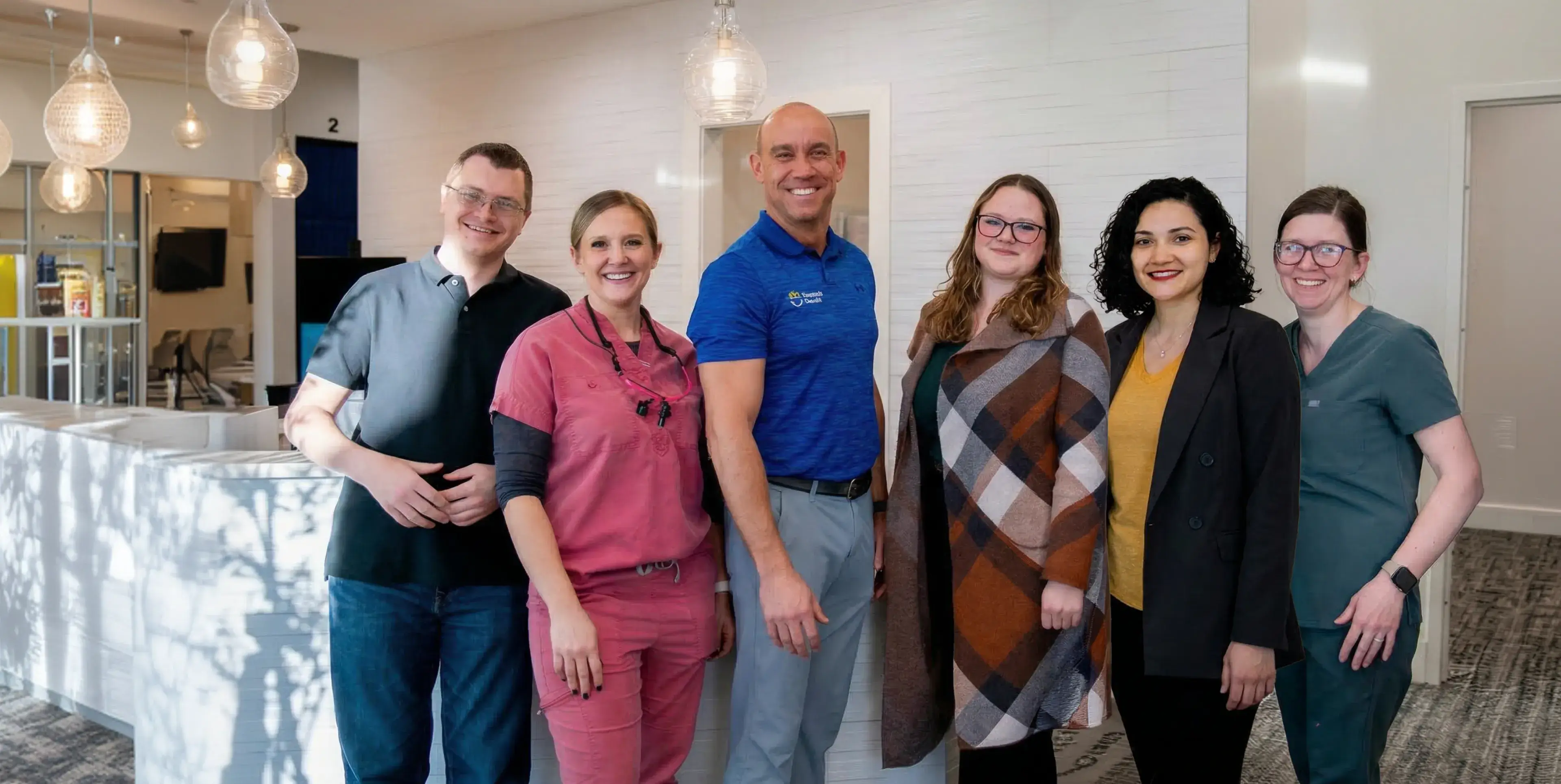 Group of six smiling dental professionals standing in an office reception area with modern lighting.