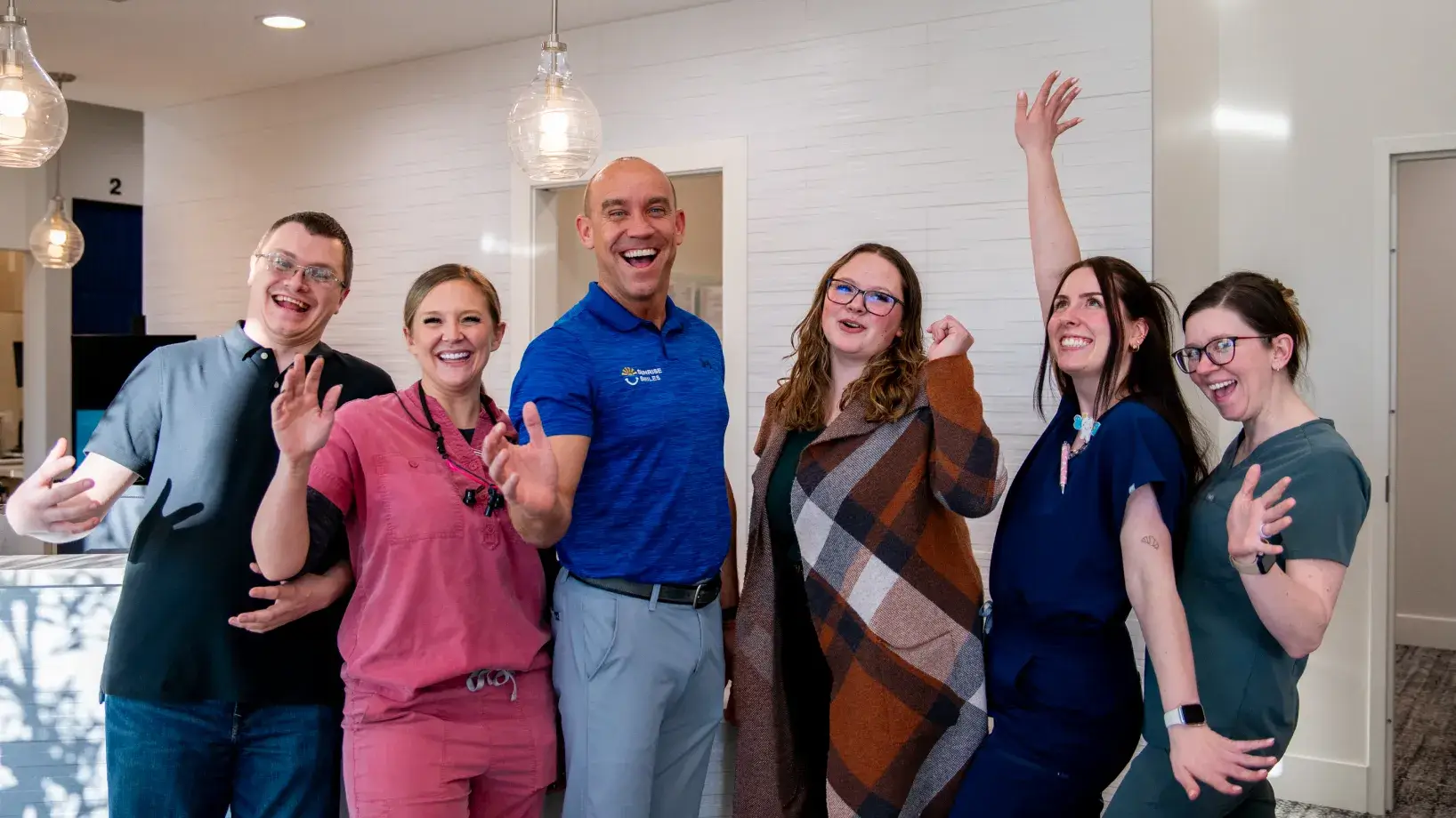 Six cheerful people standing indoors, smiling and waving at the camera.