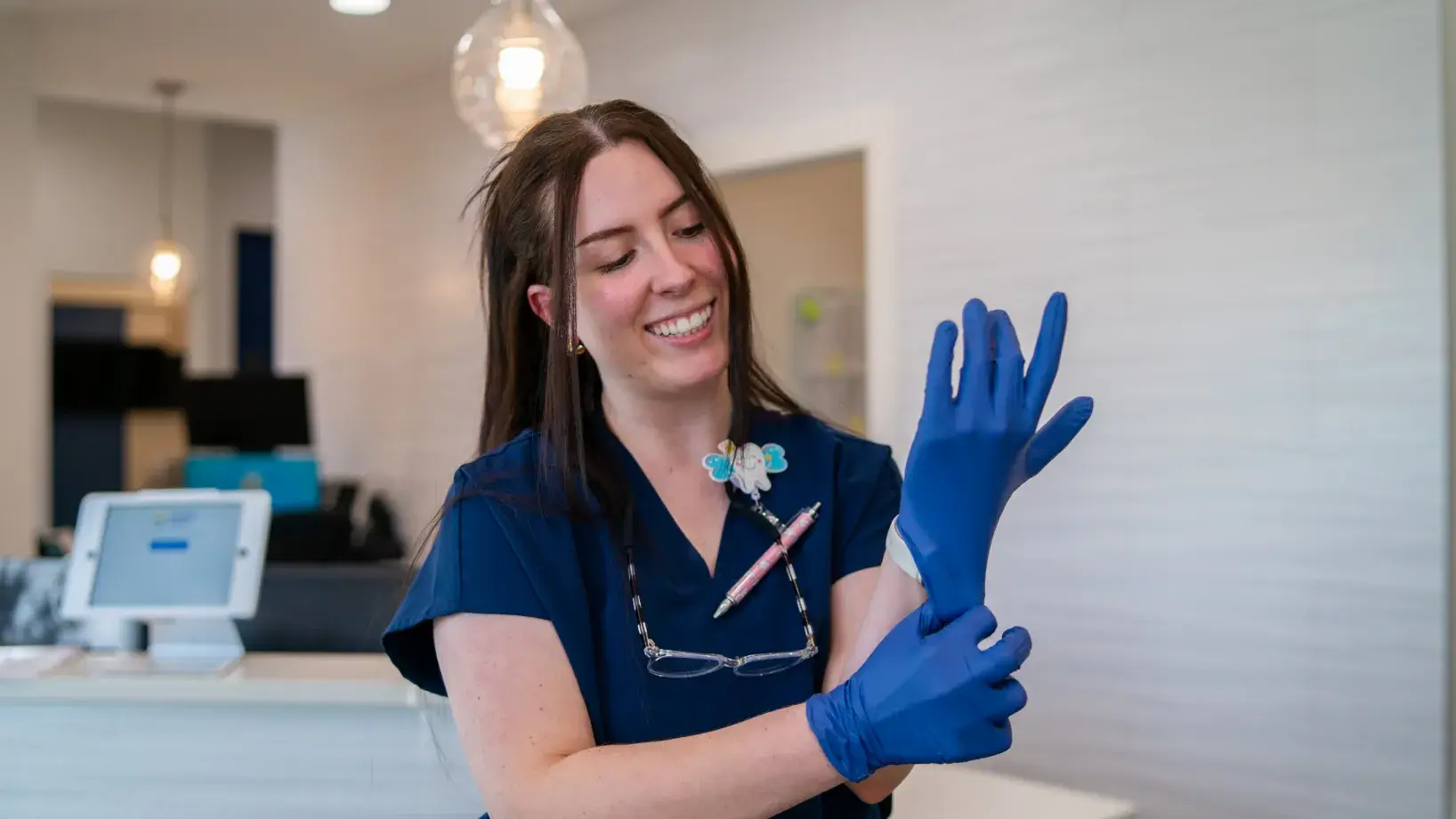 Smiling healthcare worker wearing navy scrubs putting on blue gloves in a medical office.