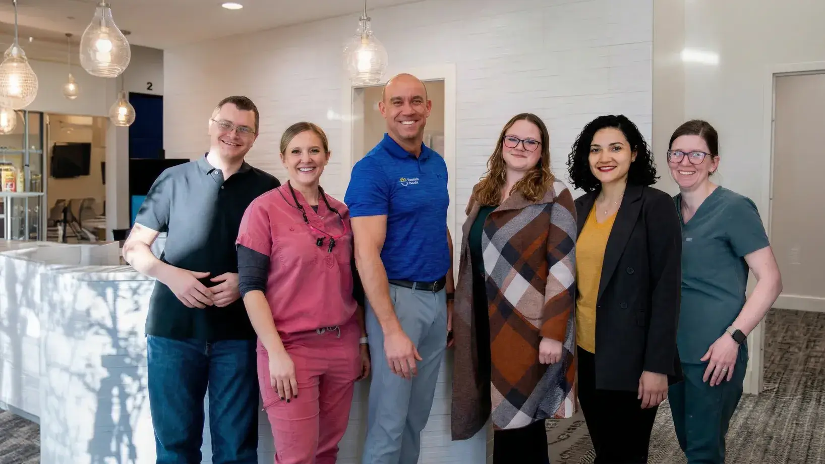 Group portrait of six smiling dental clinic staff standing indoors under modern hanging lights.