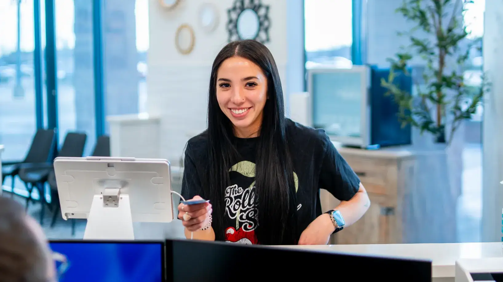 Smiling woman with long black hair holding a credit card at a white counter with a tablet stand in a modern office setting.