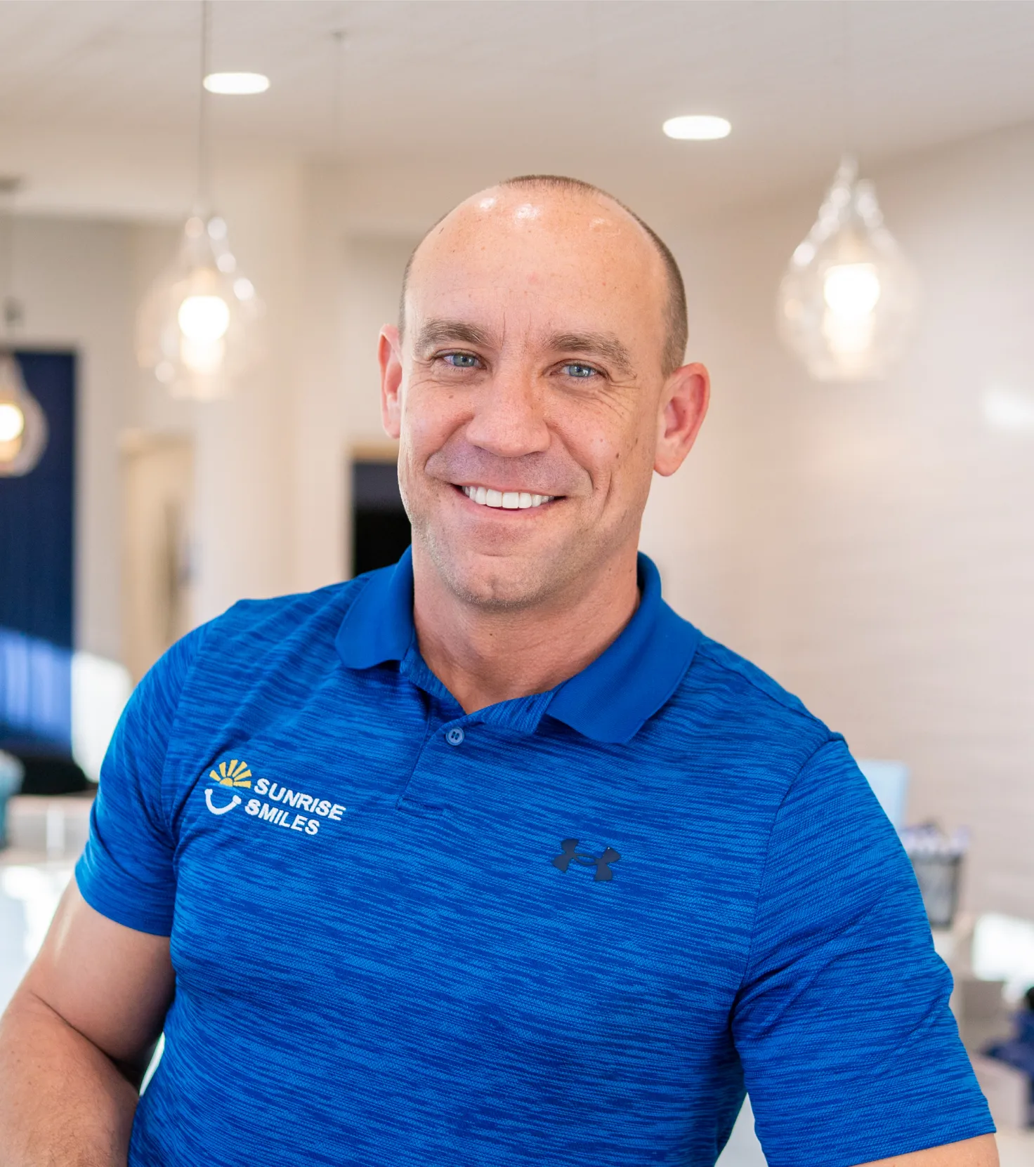 Smiling bald man in a blue Sunrise Smiles polo shirt in a bright indoor setting.