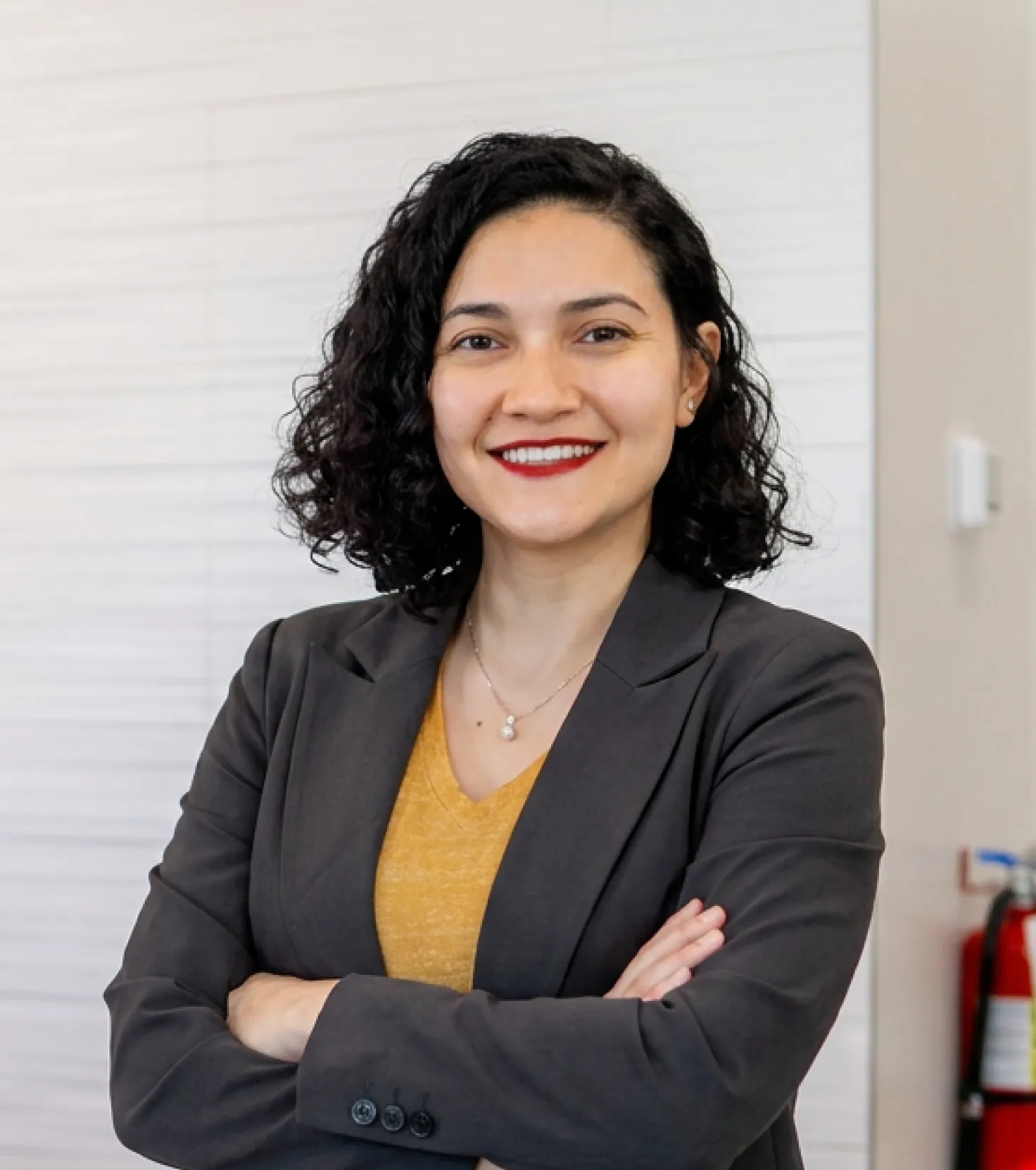 Smiling woman with curly hair wearing a dark blazer and yellow top, standing with arms crossed.