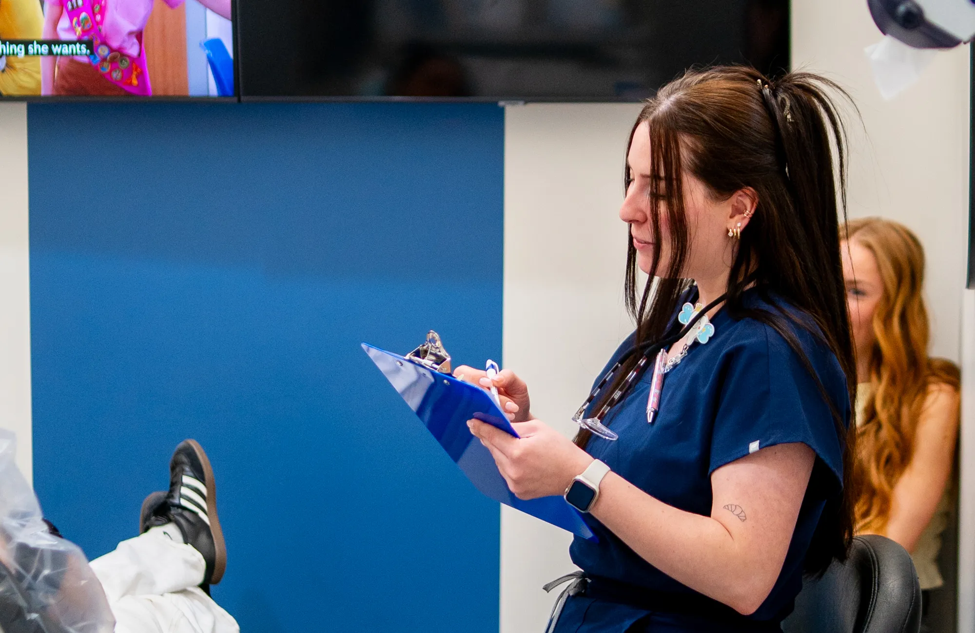 A woman in blue scrubs with a clipboard, pen, and smartwatch, seated in a medical or clinical setting.