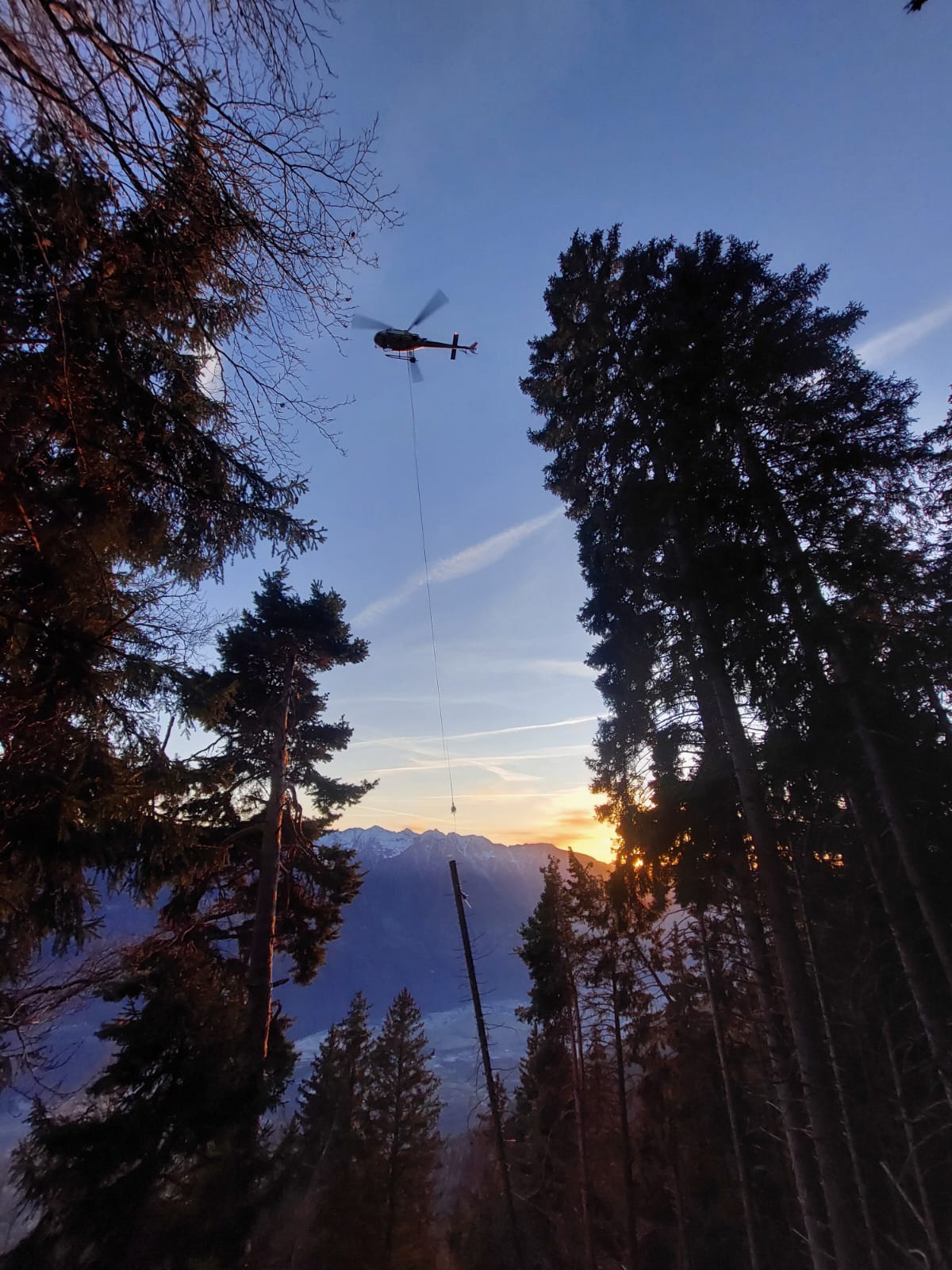 Helicopter flying over forest with mountains and sunset in the background.
