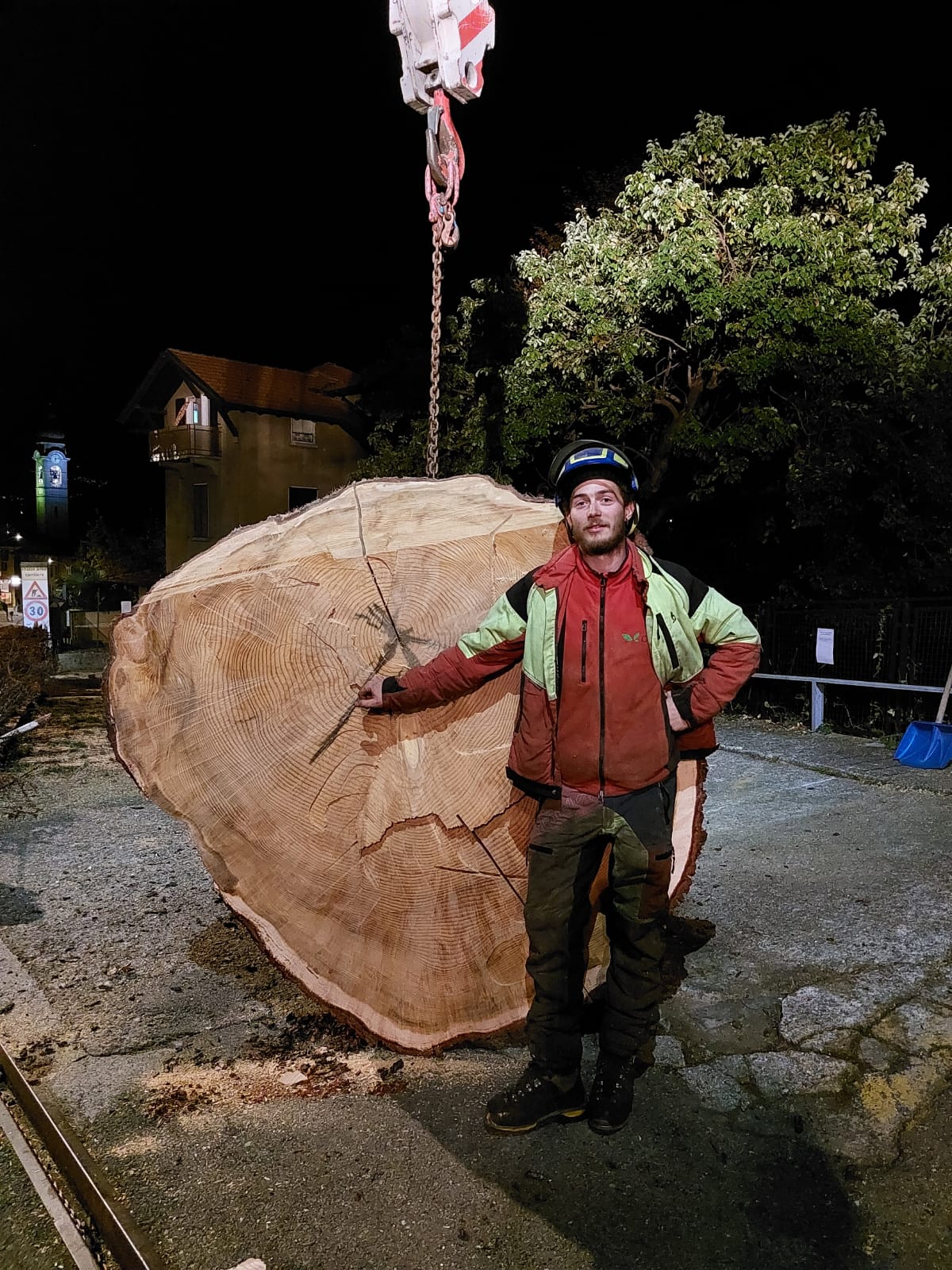 Man in safety gear standing by a large tree trunk section suspended by a crane at night.