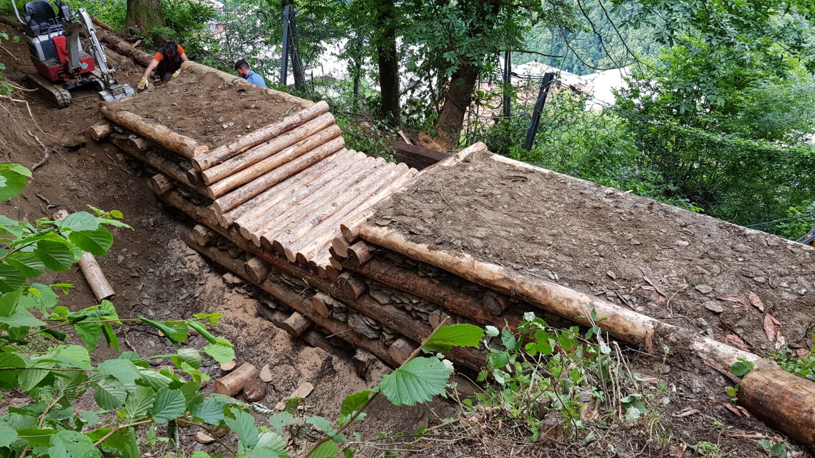 Wooden bridge construction with logs and dirt on a forested hillside, workers and equipment visible.