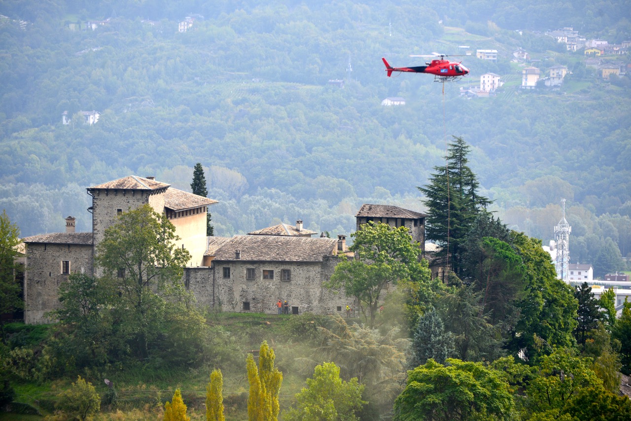 Red helicopter flying above a stone building surrounded by green trees and hills.