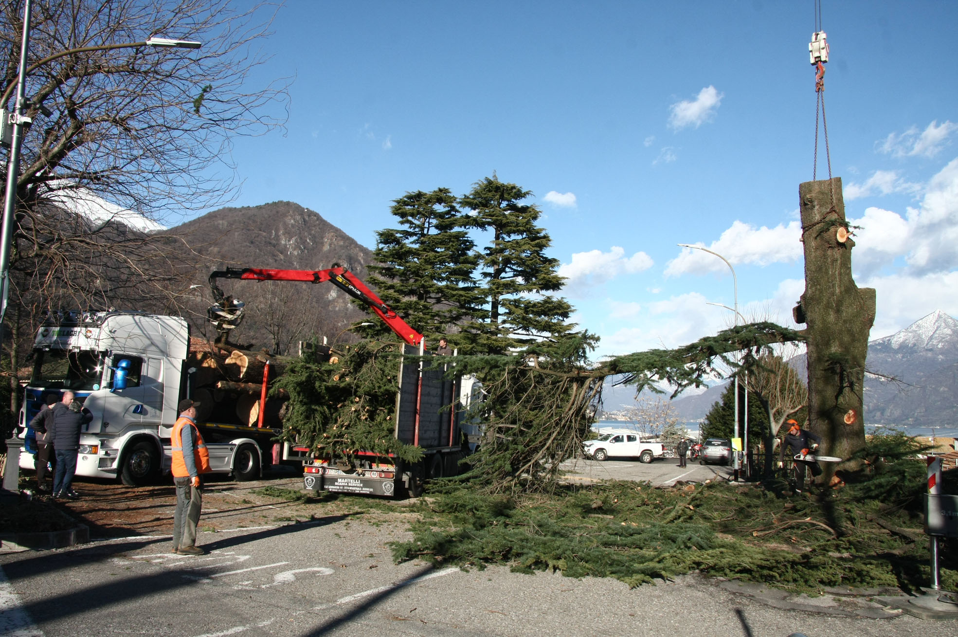 Workers cutting and loading a large tree trunk with crane and truck on a mountain road under blue sky.