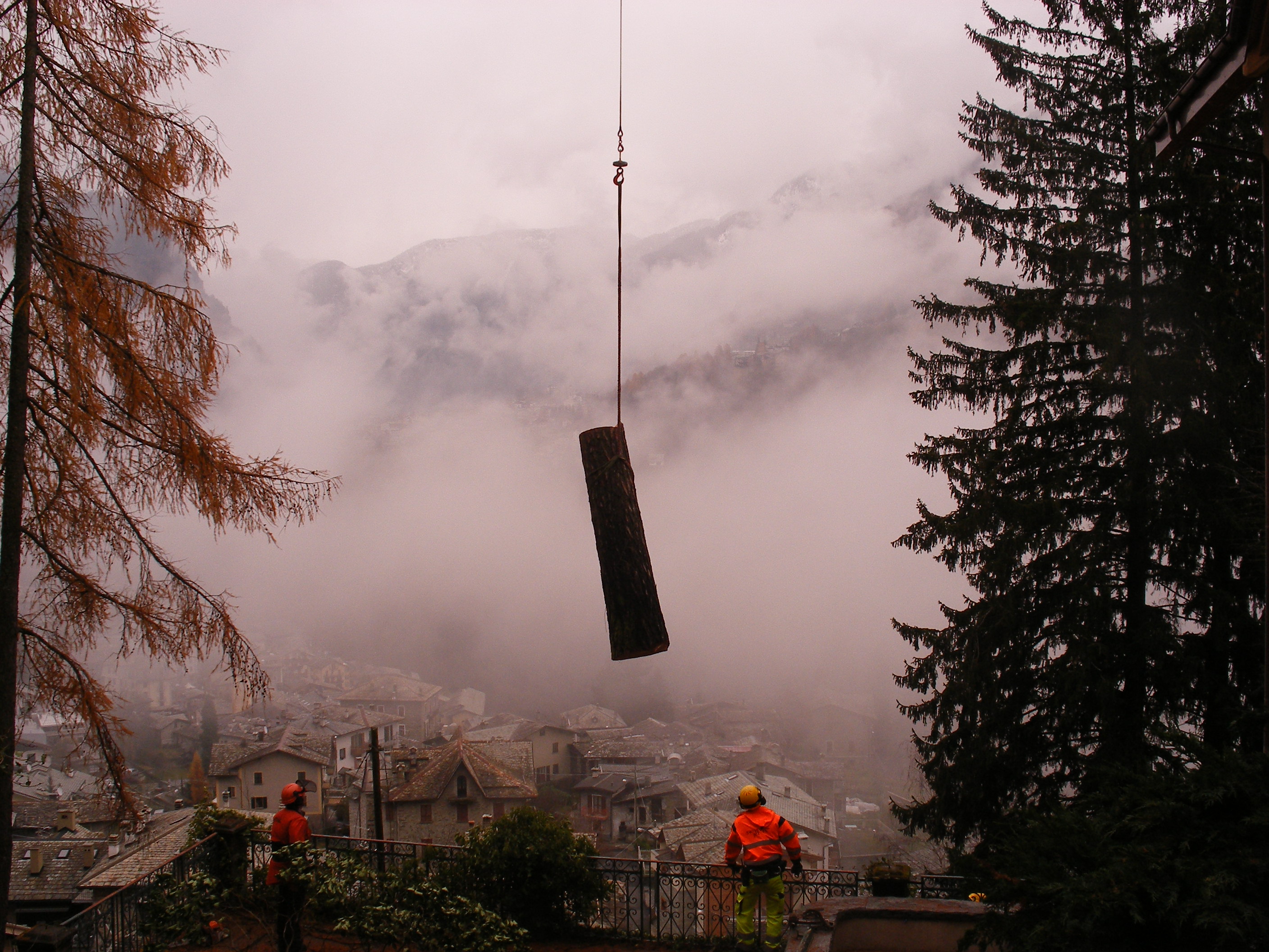 Workers in safety gear guiding a log suspended by a crane over a foggy mountain village.