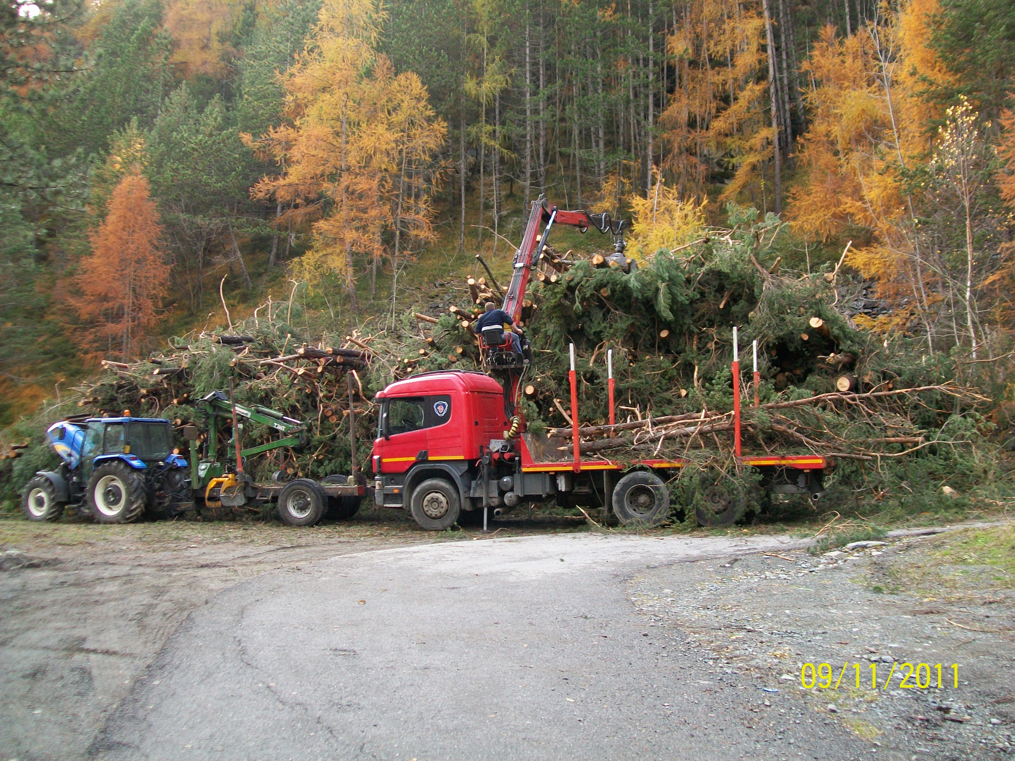 Red logging truck with crane loads cut tree branches in autumn forest clearing with tractor nearby.