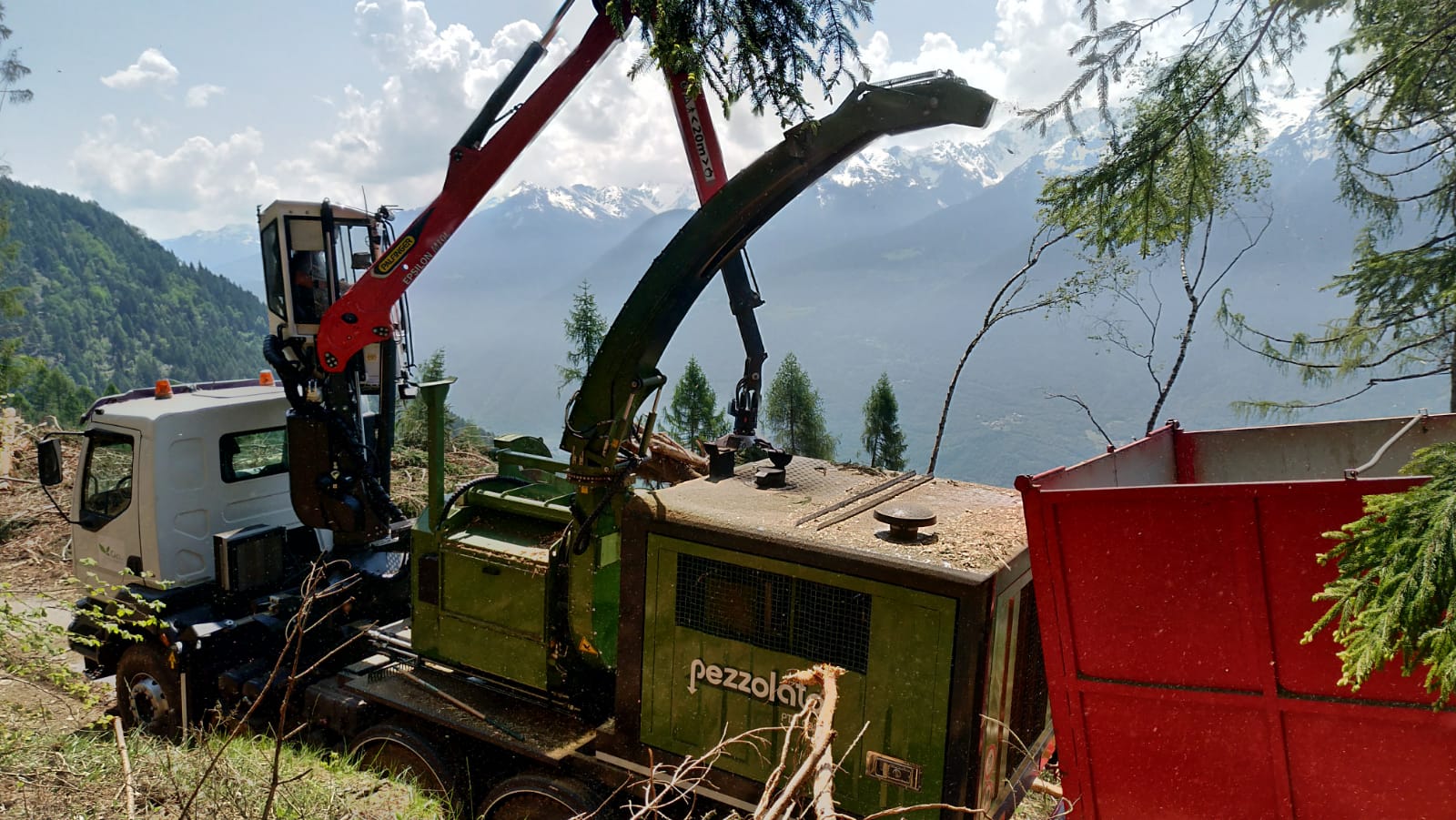 Logging truck with Pezzolato chipper in a mountainous forest with snow-capped peaks in the background.