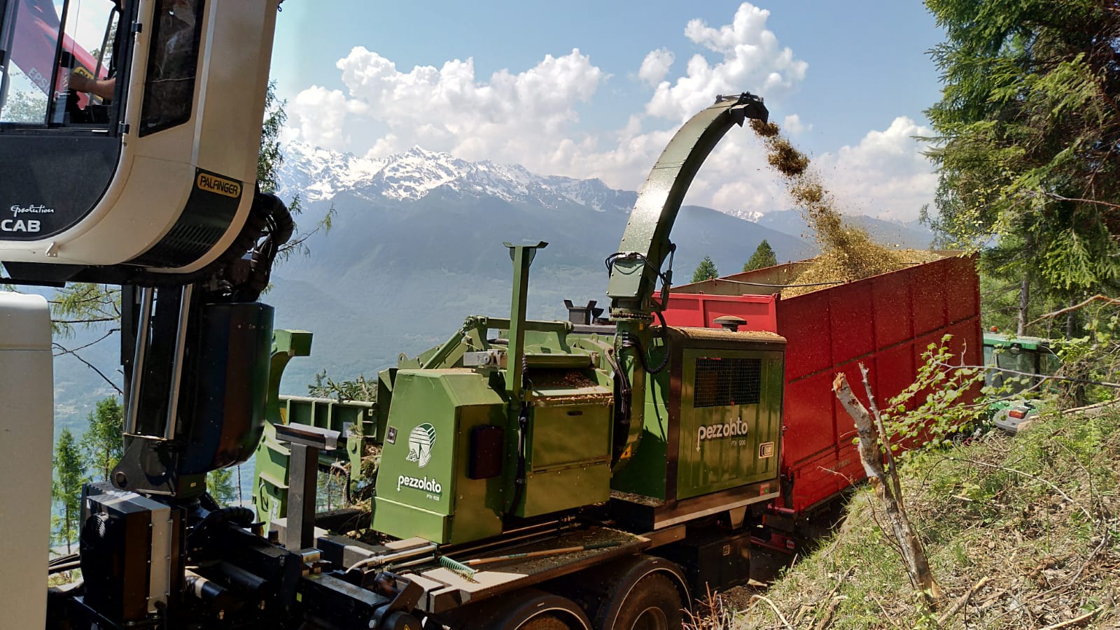Green wood chipper machine unloading chips into a red trailer on a mountain slope with snowy peaks behind.