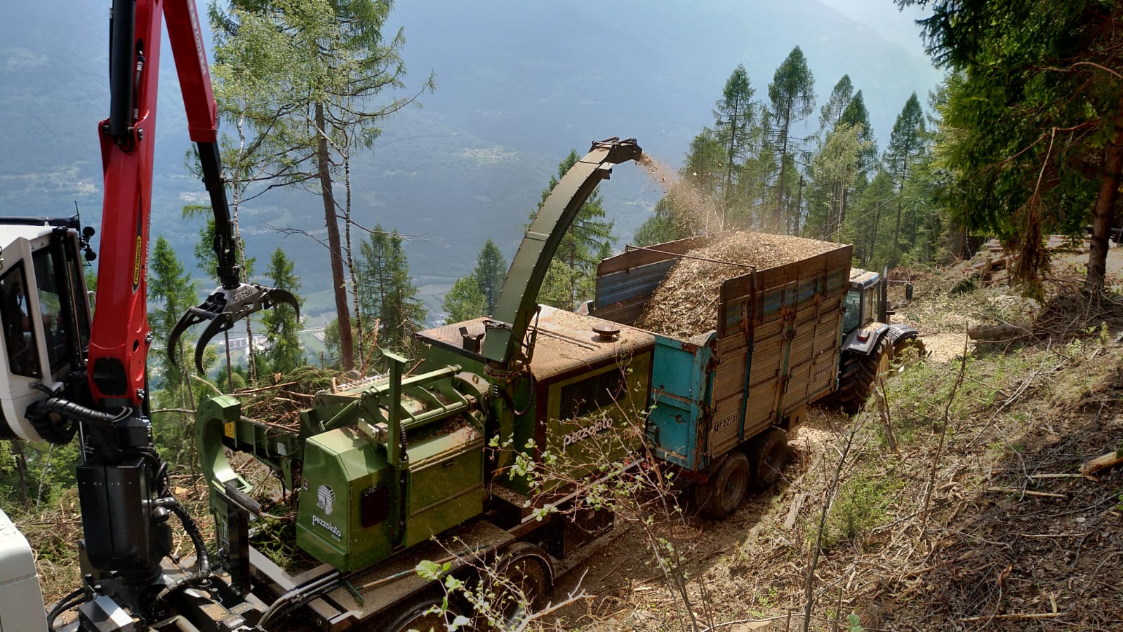 Forest machine shredding branches into wood chips loaded into a large tractor trailer on a hillside.