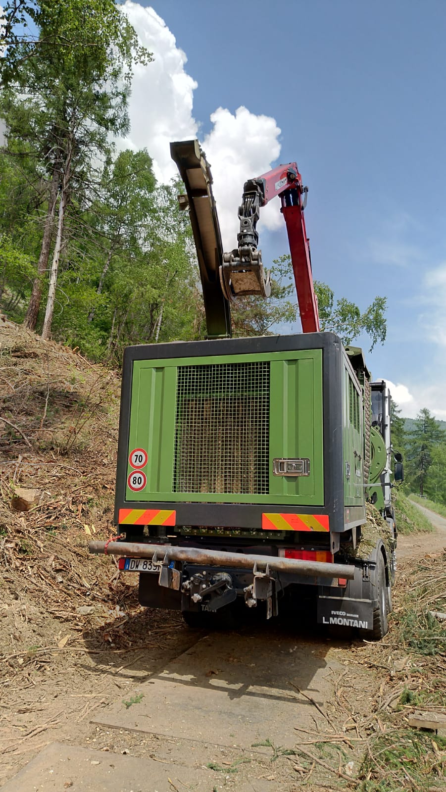 Rear view of green truck with red crane arm on dirt road in wooded area under blue sky.