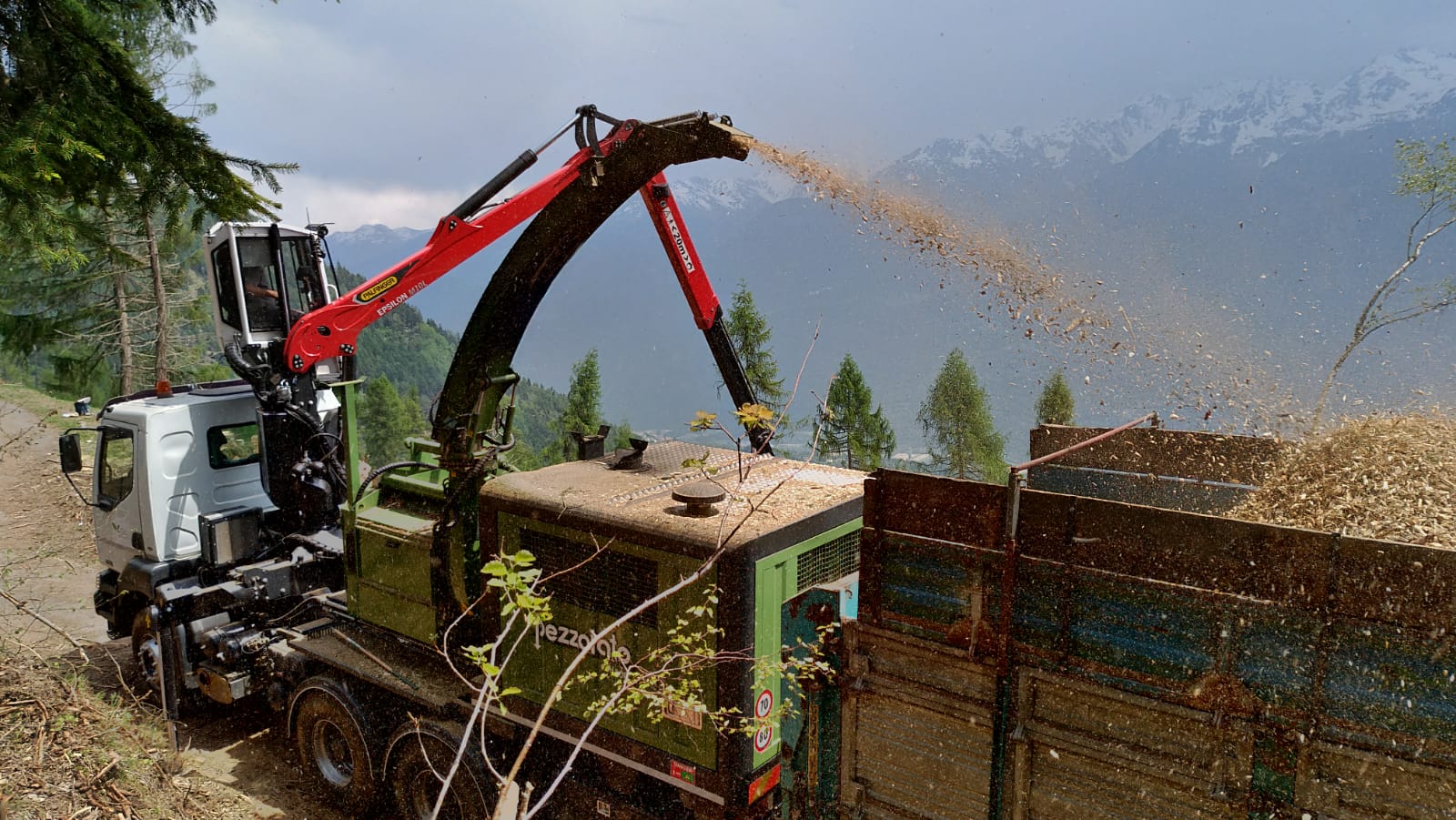 Wood chipper truck shredding branches and ejecting wood chips into a trailer with forest and mountains behind.