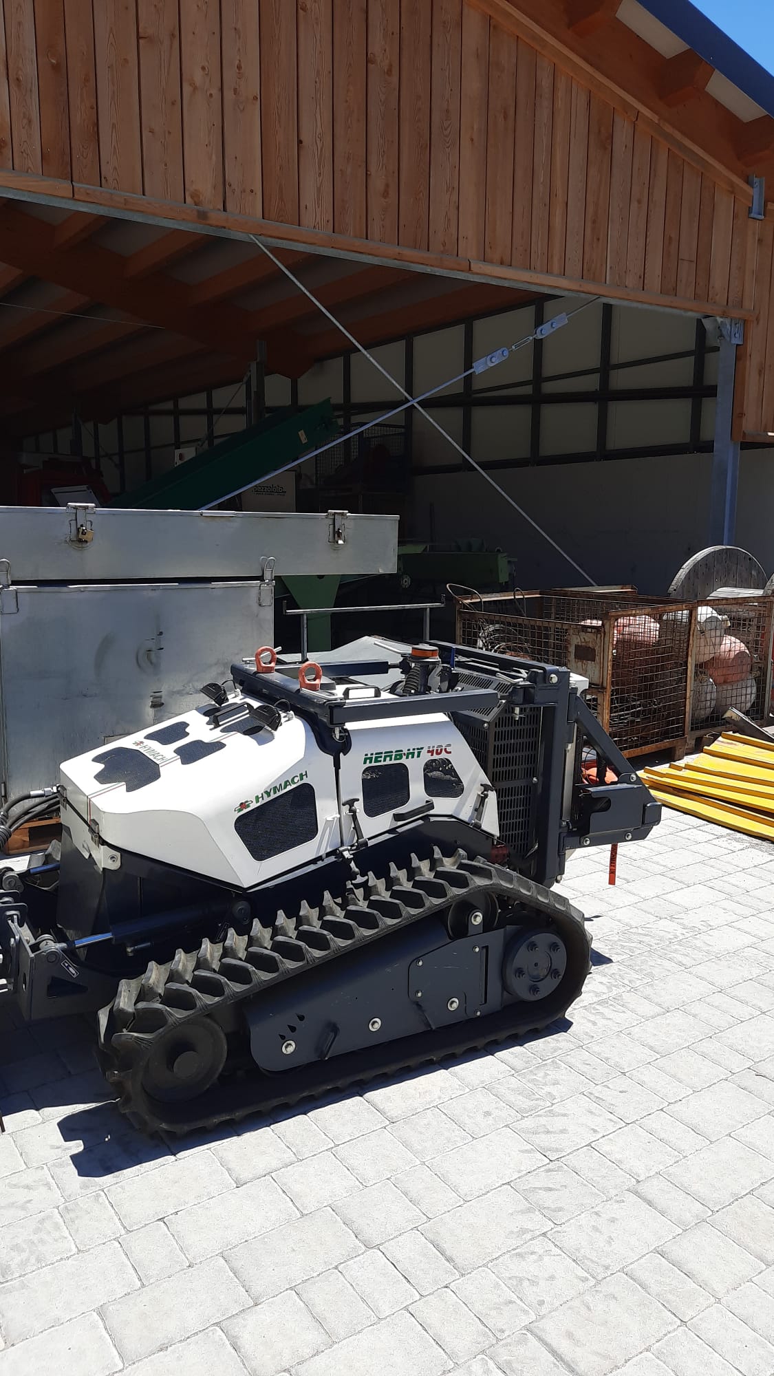 Tracked robotic vehicle labeled HYMACH HERBAT 40C parked on gray paved ground by a wooden building.