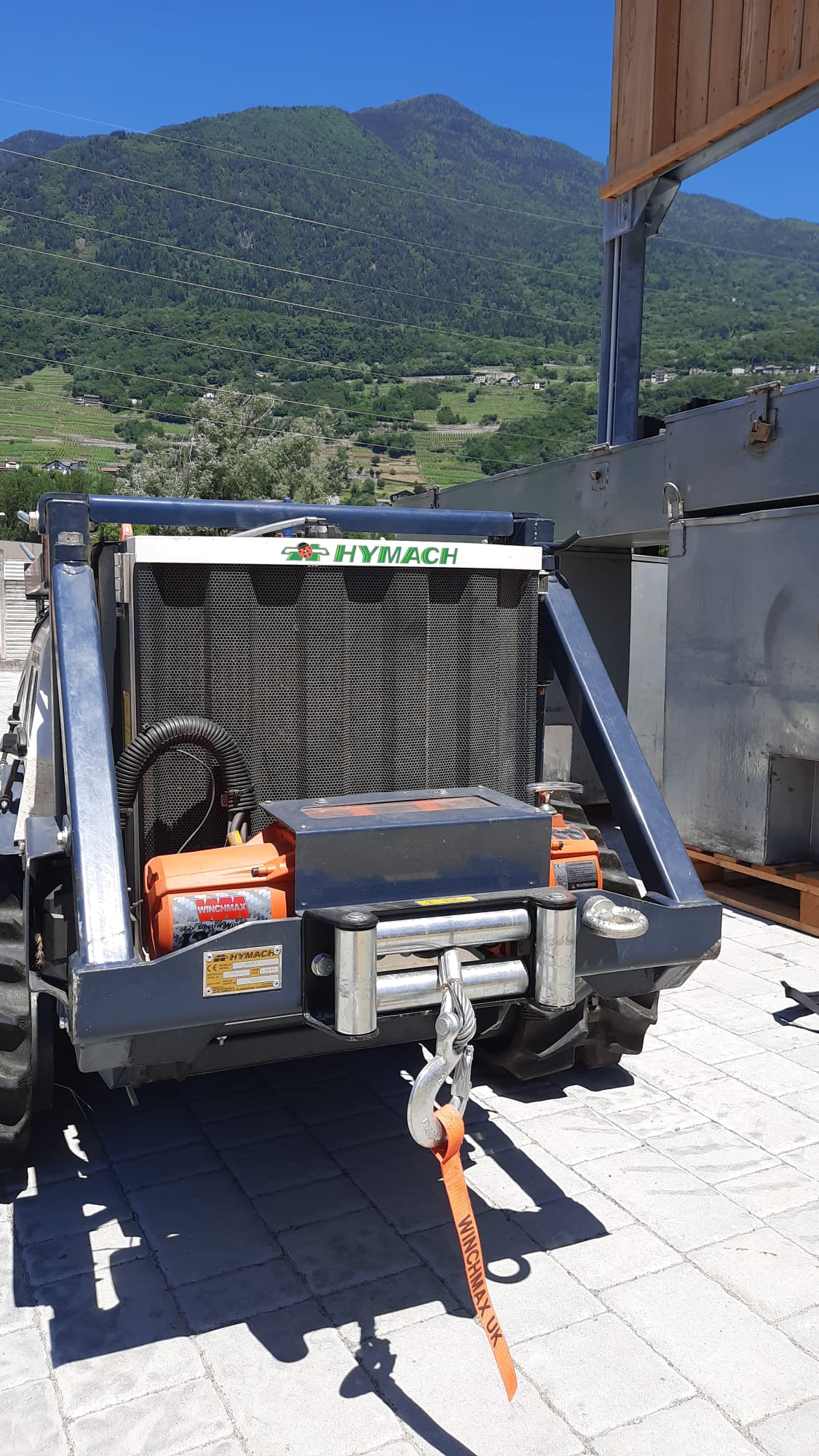 Front view of a black and orange HY MACH winch attached to a tractor with green mountains in background.