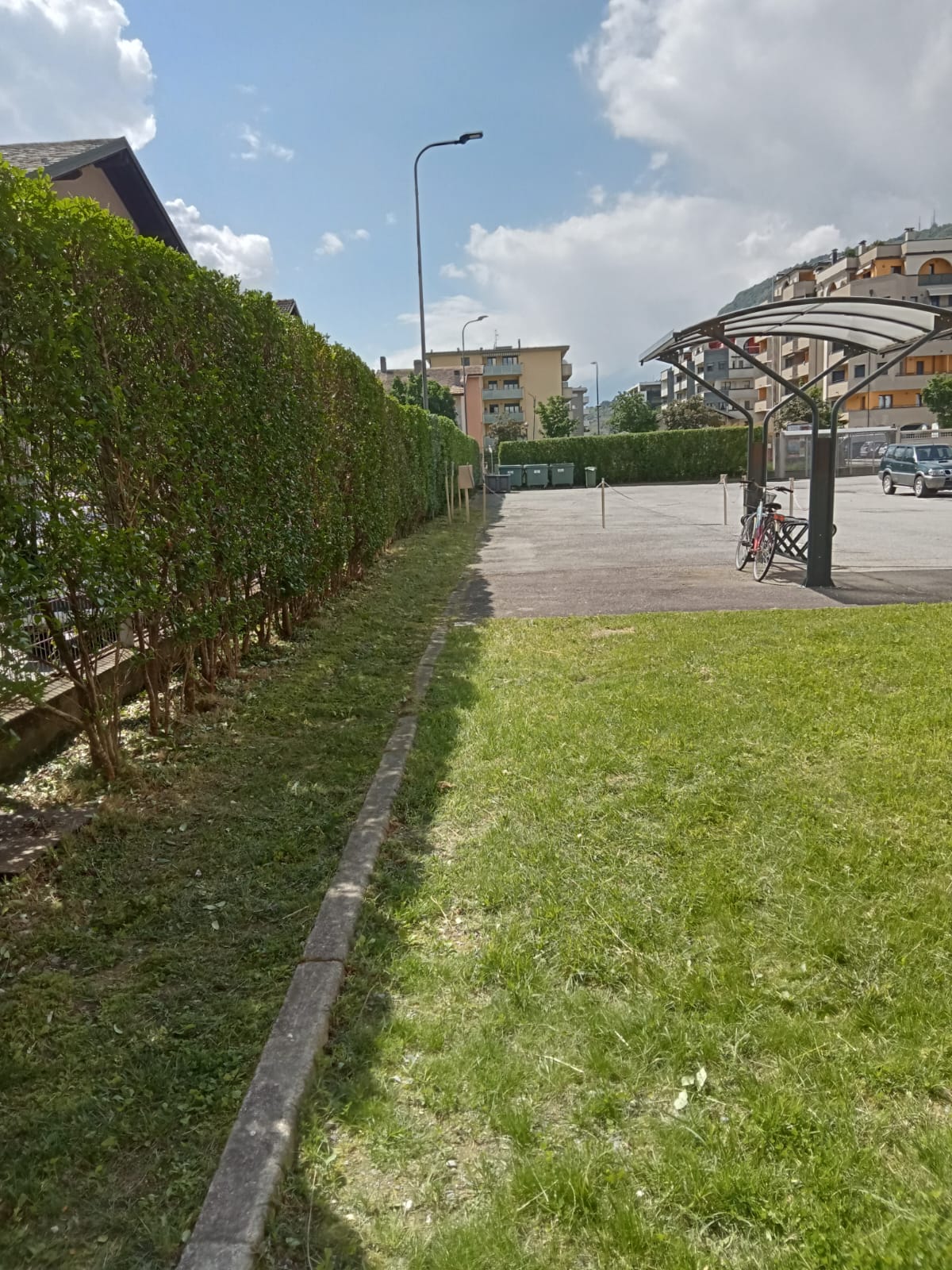 Green lawn and hedge along a sidewalk, parking area with bike shelter and residential buildings in background.