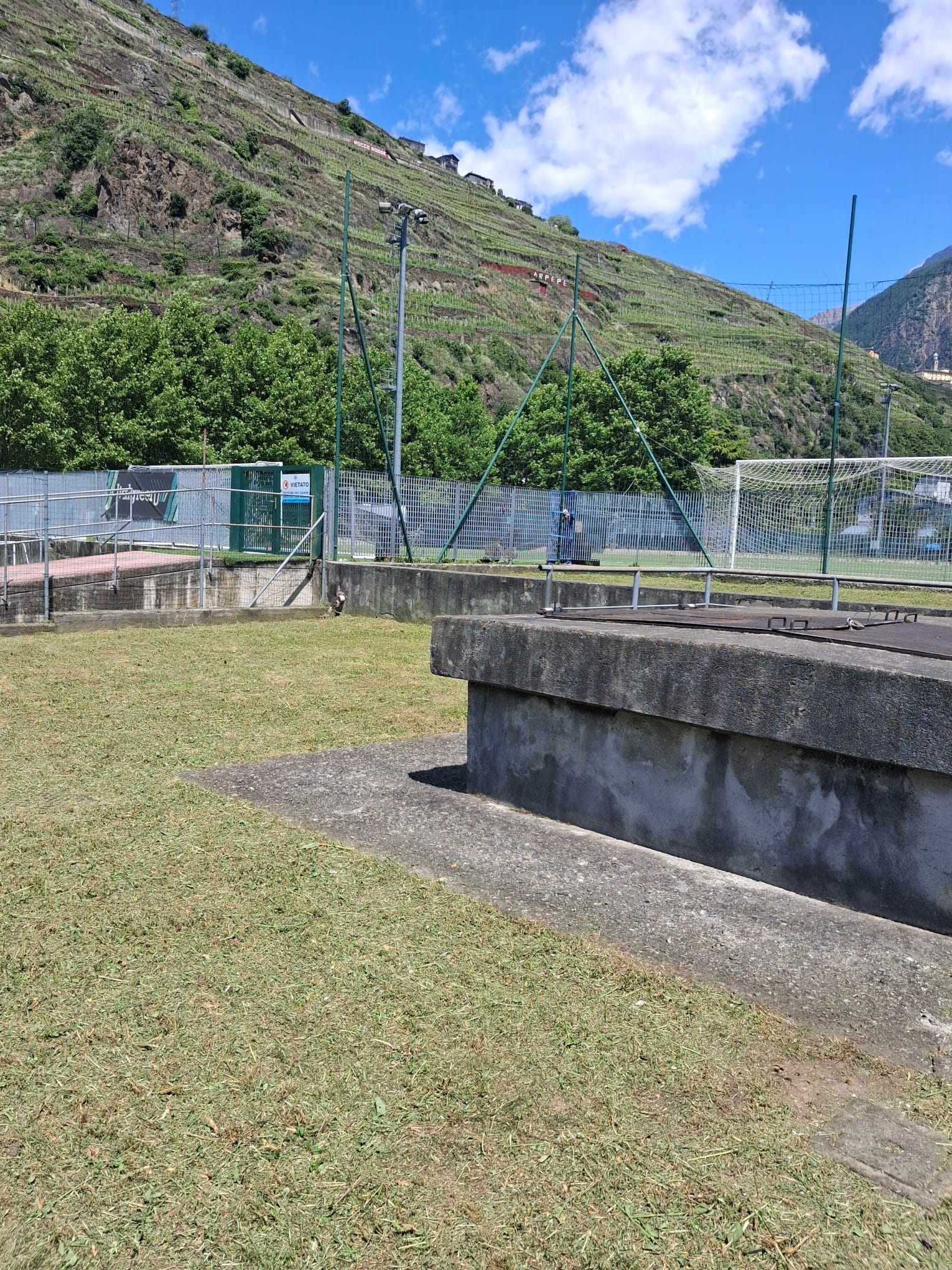 Grassy outdoor area with concrete platform, fenced soccer goal, trees, and terraced hillside under blue sky.