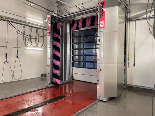 Binnenkant van een automatische wasstraat met rode natte vloer en grote draaiende borstels in zwart en roze.