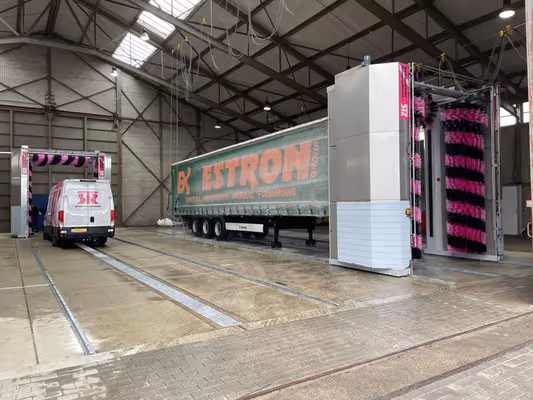 Binnenkant van een wasstraat met een witte bestelwagen aan de linkerkant en een grote vrachtwagen met het logo 'EK ESTRON' in het midden, omgeven door automatische wasborstels.