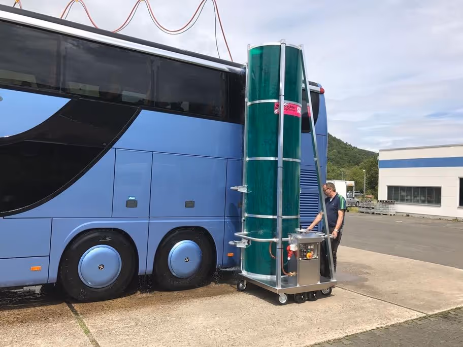Man bedient een grote, groene automatische wasborstel die een blauwe touringbus schoonmaakt.