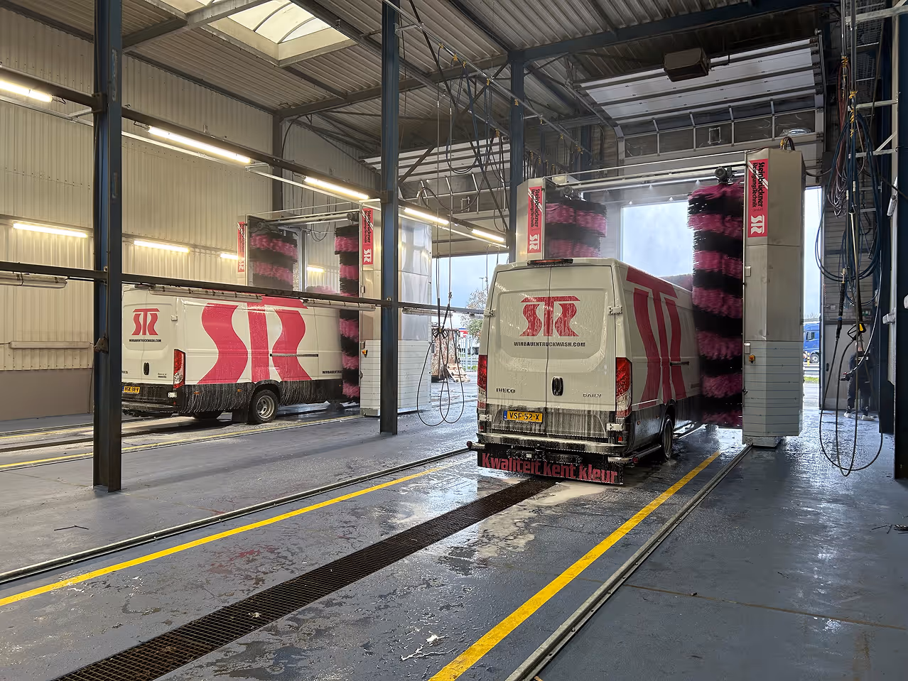 Een witte bestelwagen met rode SB-logo's wordt schoongemaakt in een automatische wasstraat met roze-zwart borstels.