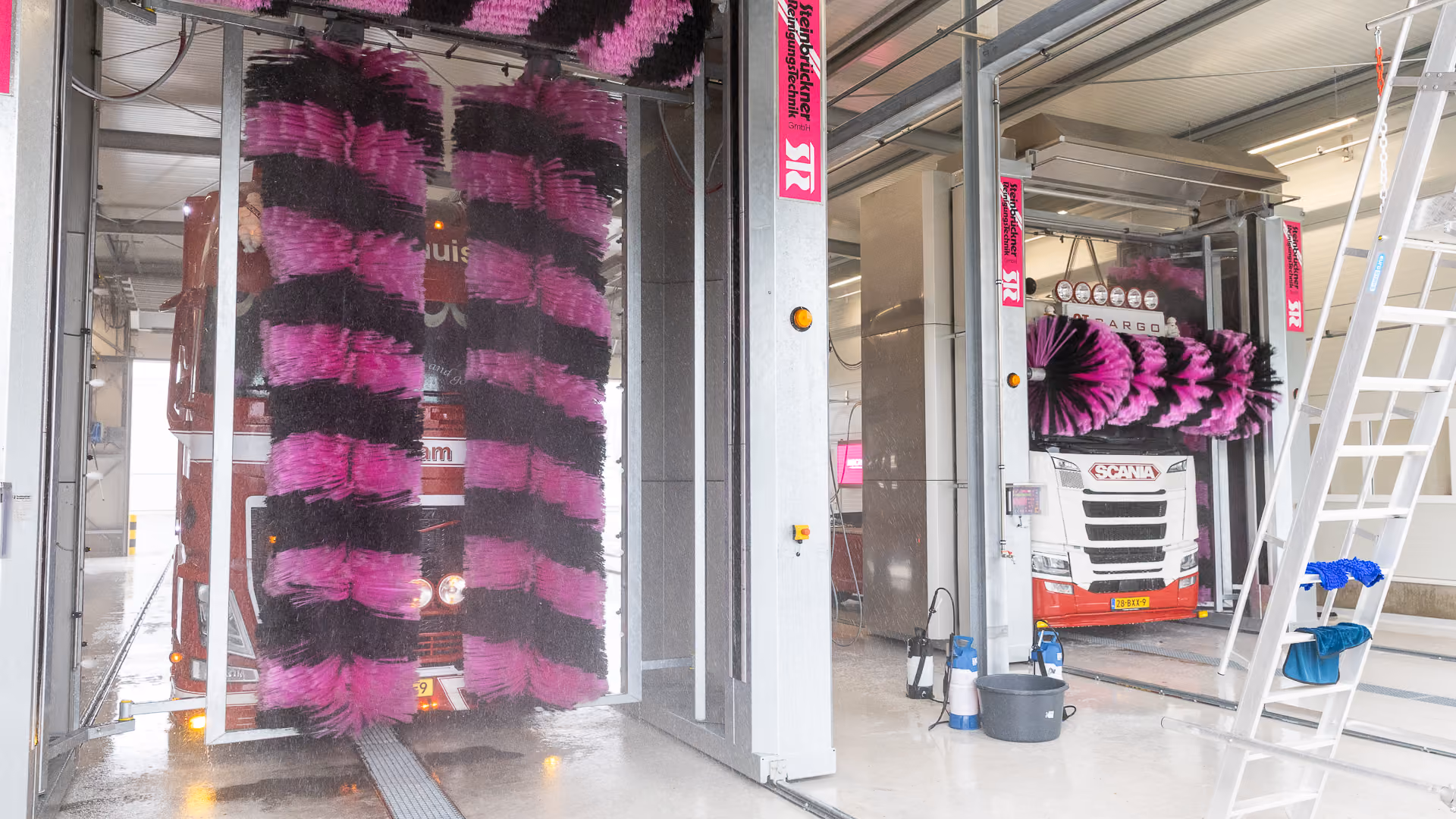Twee vrachtwagens in een automatische wasstraat met grote roterende borstels in zwarte en roze kleuren die de wagens schoonmaken in een overdekte wasfaciliteit.