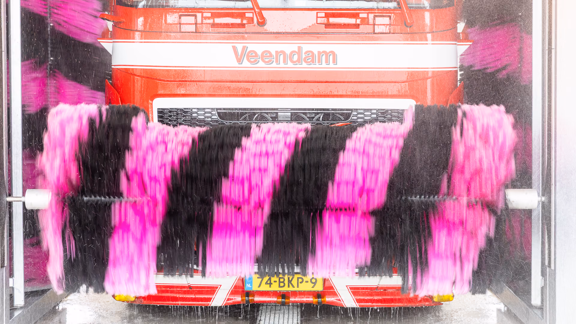 Rood witte vrachtwagen met naam Veendam krijgt wasbeurt in automatische wasstraat met draaiende roze en zwarte borstels vooraan.