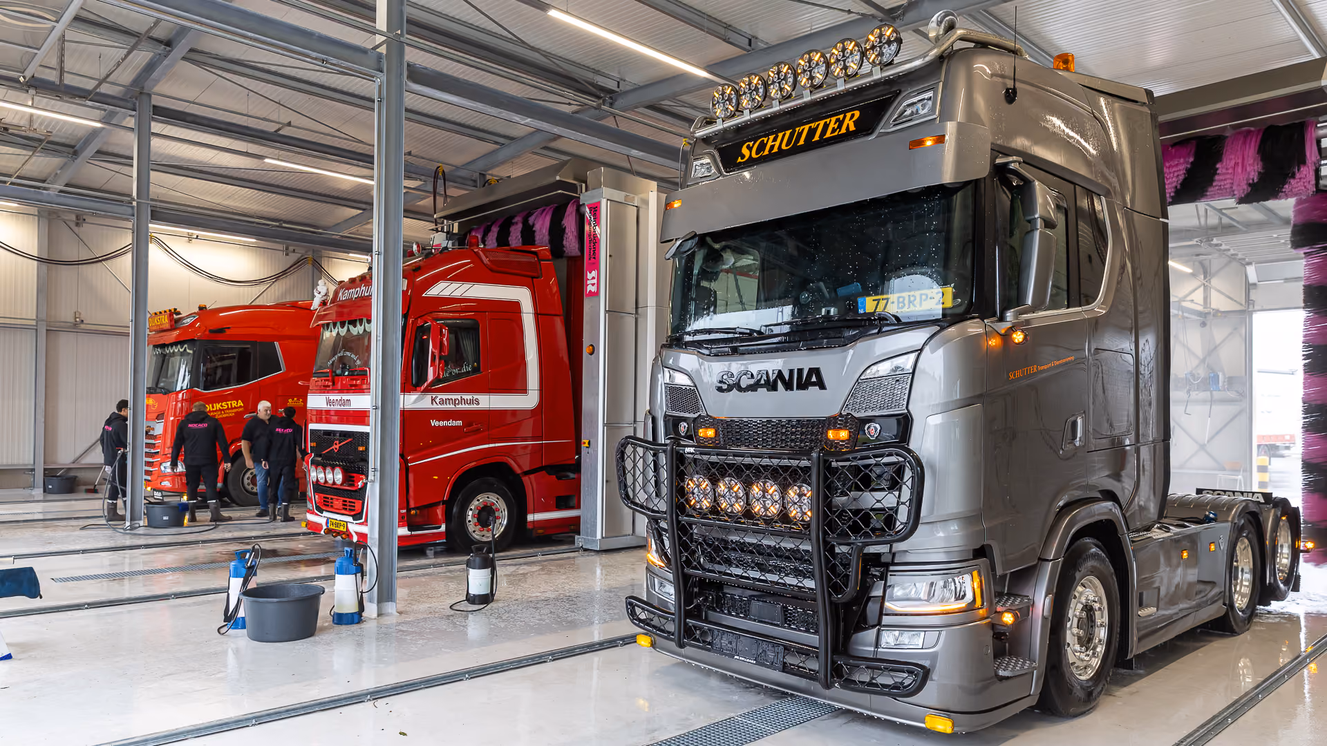 Een moderne grijze Scania vrachtwagen met een zwaar beschermrek en extra verlichting geparkeerd in een grote, schone wasruimte naast een rode vrachtwagen terwijl mensen werkzaamheden uitvoeren.
