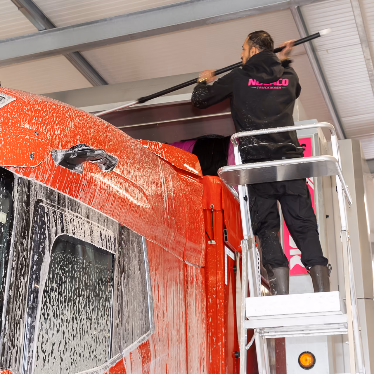 Een persoon staat op een metalen trap en maakt een rood vrachtwagenhuis schoon met een lange borstel, bedekt met schuim, in een binnenruimte.